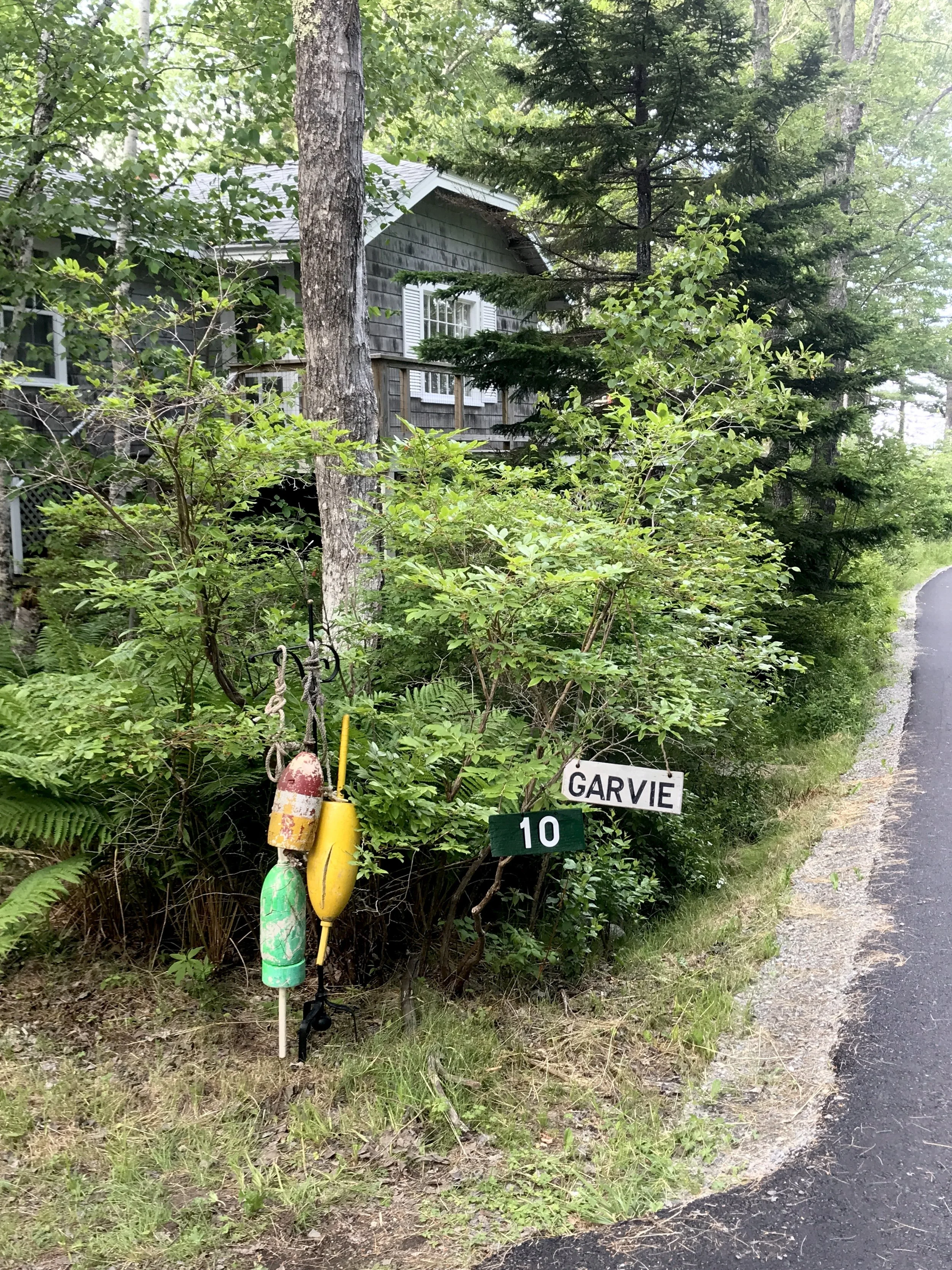 A wooded roadside scene with a house partially visible in the background among trees. There are buoys hanging from a small stand with signs that read 'GARVIE' and the number '10'.