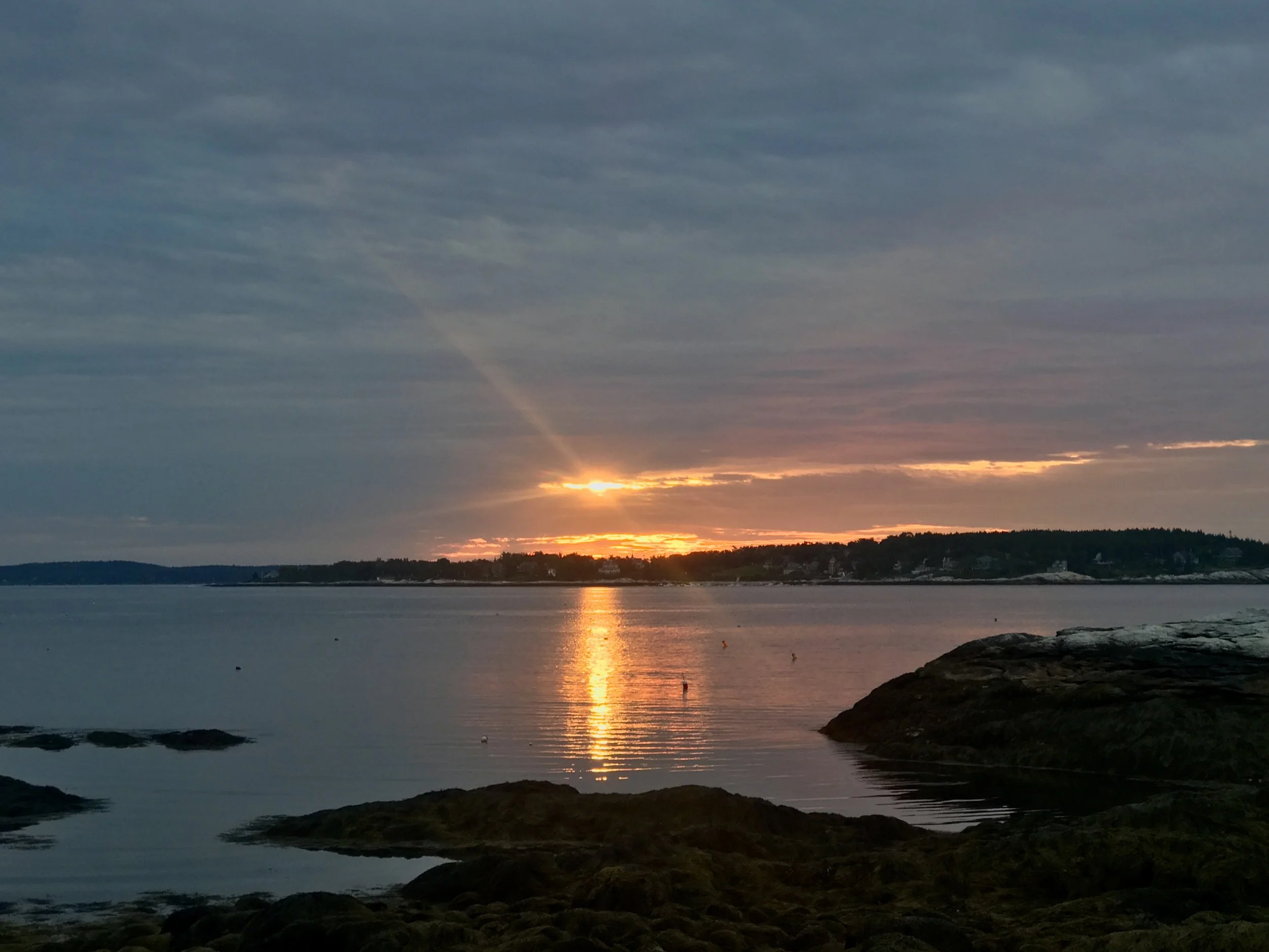 Sunrise over a calm body of water with rocky shoreline and distant land, sky partly cloudy with rays of sunlight