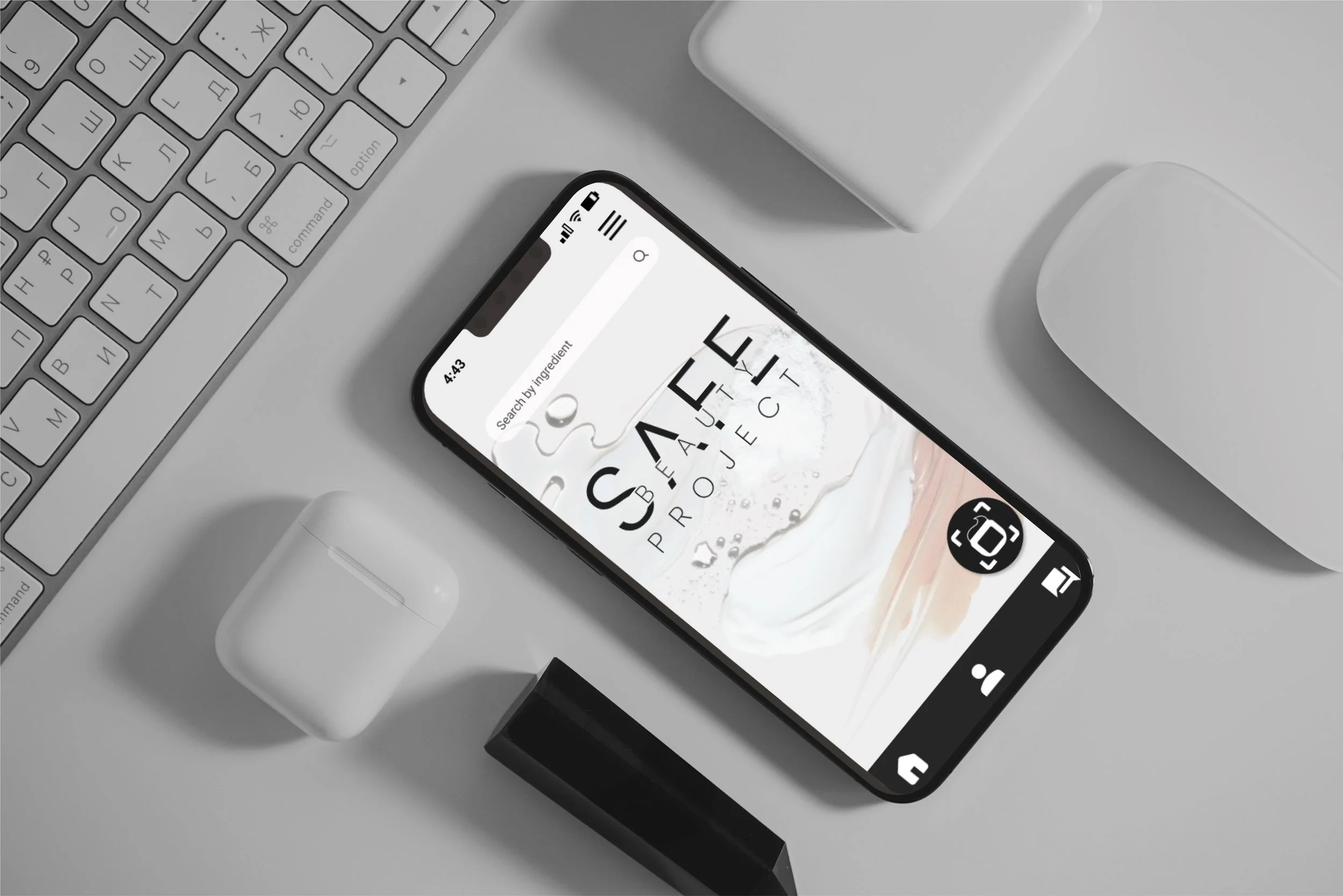 A smartphone on a desk displaying an app with the text 'SAFE BEAUTY PROJECT' on its screen, surrounded by wireless earbuds in a case, a lip balm, a computer keyboard, a white mouse, and a rectangular container.