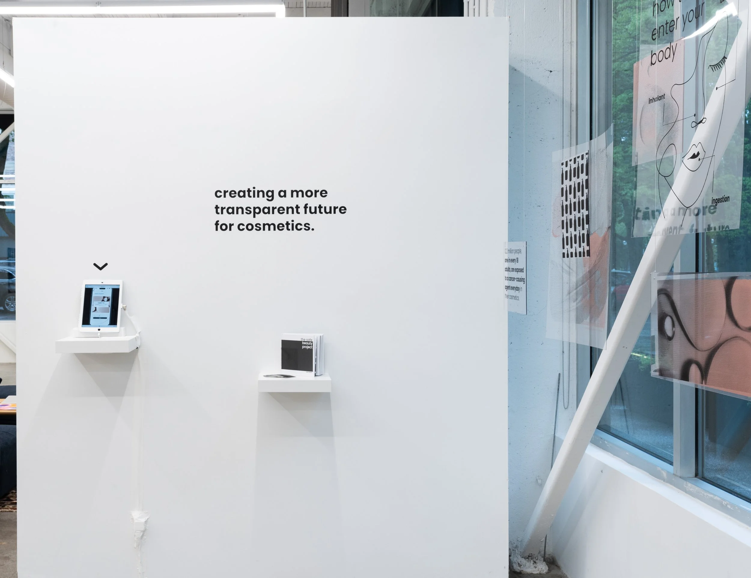 White gallery wall with black text that reads, "creating a more transparent future for cosmetics," with two small display shelves holding digital devices and printed materials.