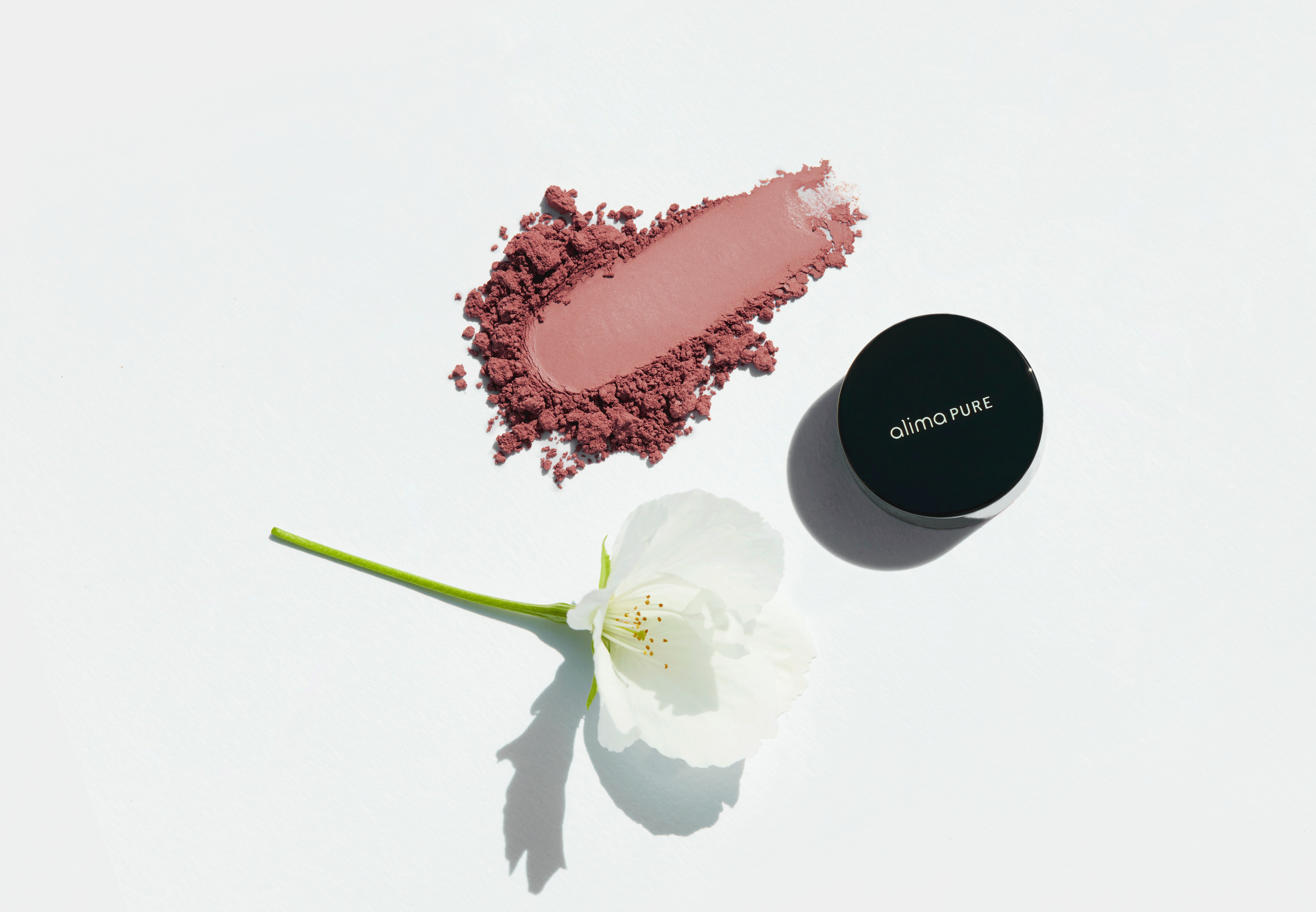 A black container labeled 'alima PURE' next to a pink powdered makeup product and a white flower on a white background.