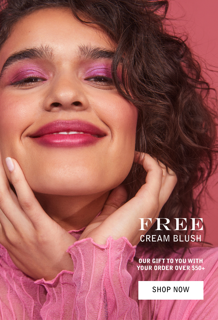 Close-up of a smiling woman with curly brown hair, pink eyeshadow, pink lipstick, and a pink ruffled long sleeve top, against a pink background.