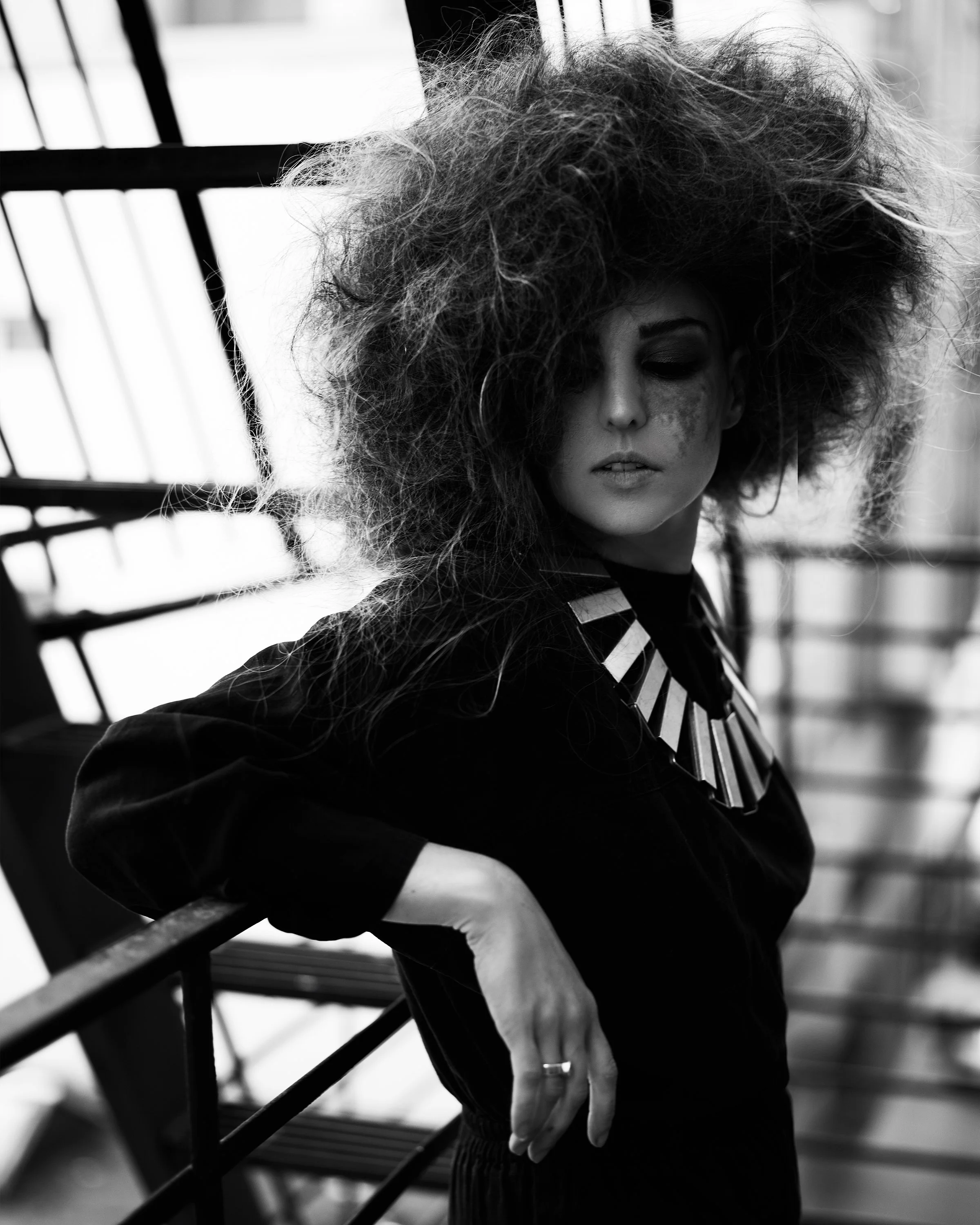 A woman with wild, voluminous hair leaning on a railing in an industrial setting, wearing a dark outfit and a chunky statement necklace, in black and white.