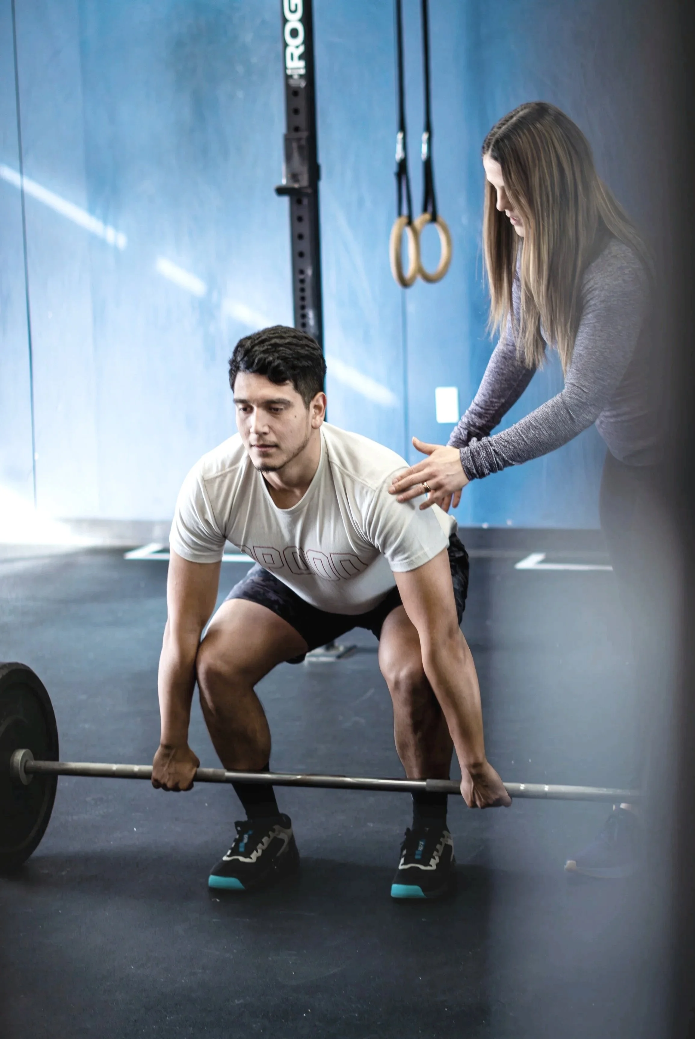 A man is lifting a barbell with weights, while a woman is assisting or coaching him in a gym.