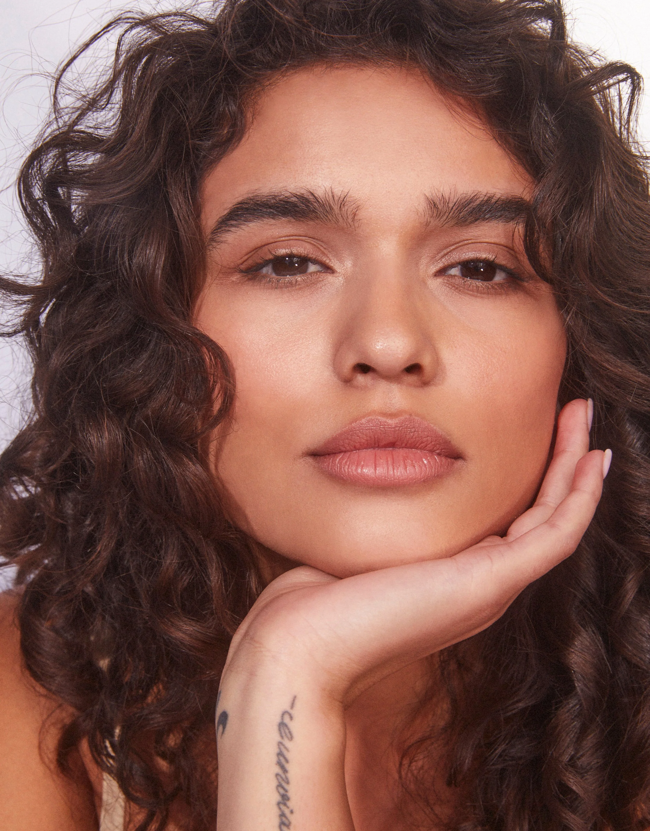 A woman with curly brown hair resting her chin on her hand, looking directly at the camera. She has natural makeup, full lips, and a tattoo on her wrist that reads 'Confidence.'