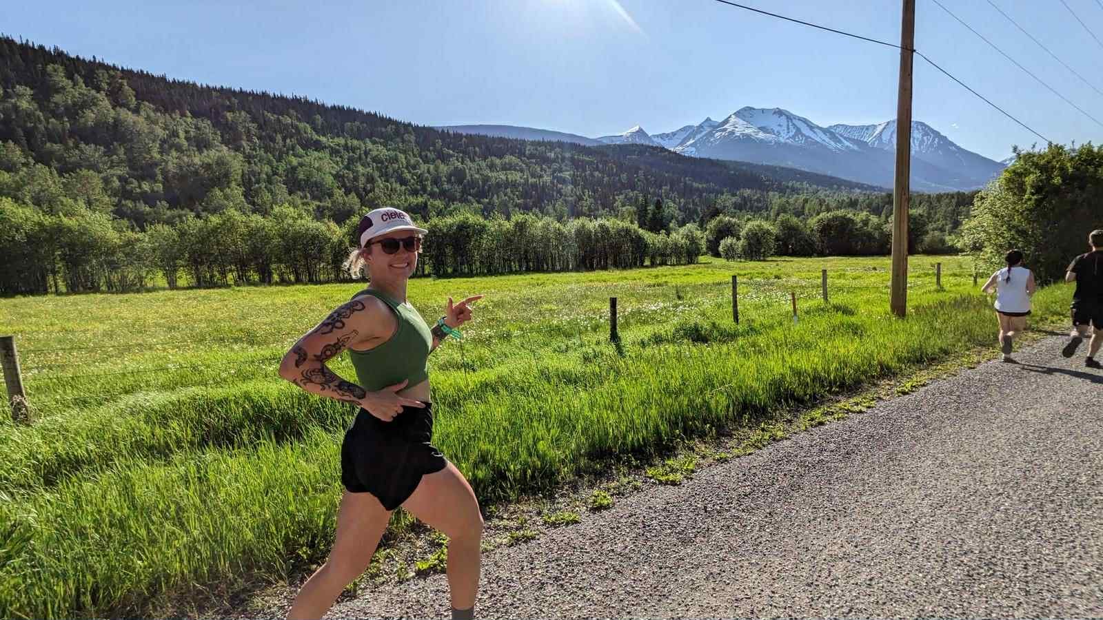 A woman in athletic clothing and sunglasses in a running pose on a gravel trail in a scenic mountain landscape with green fields, trees, and snow-capped mountains in the background.