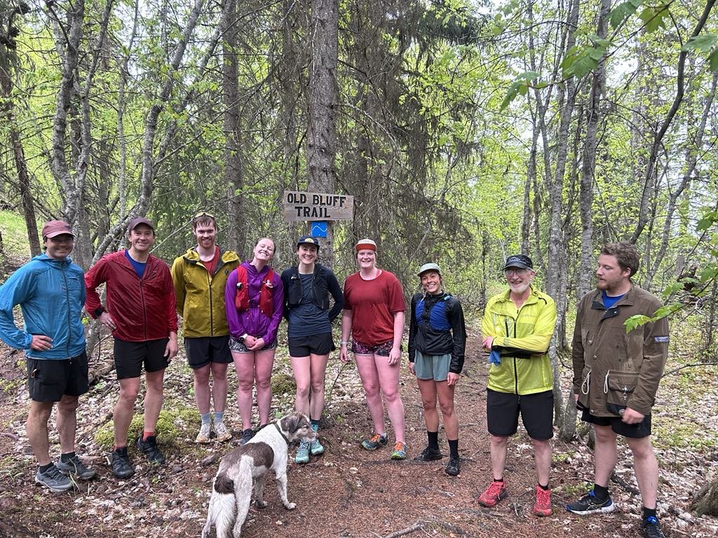 Group of nine hikers and a dog standing on a trail in the woods, with a sign that reads 'Old Bluff Trail' in the background.