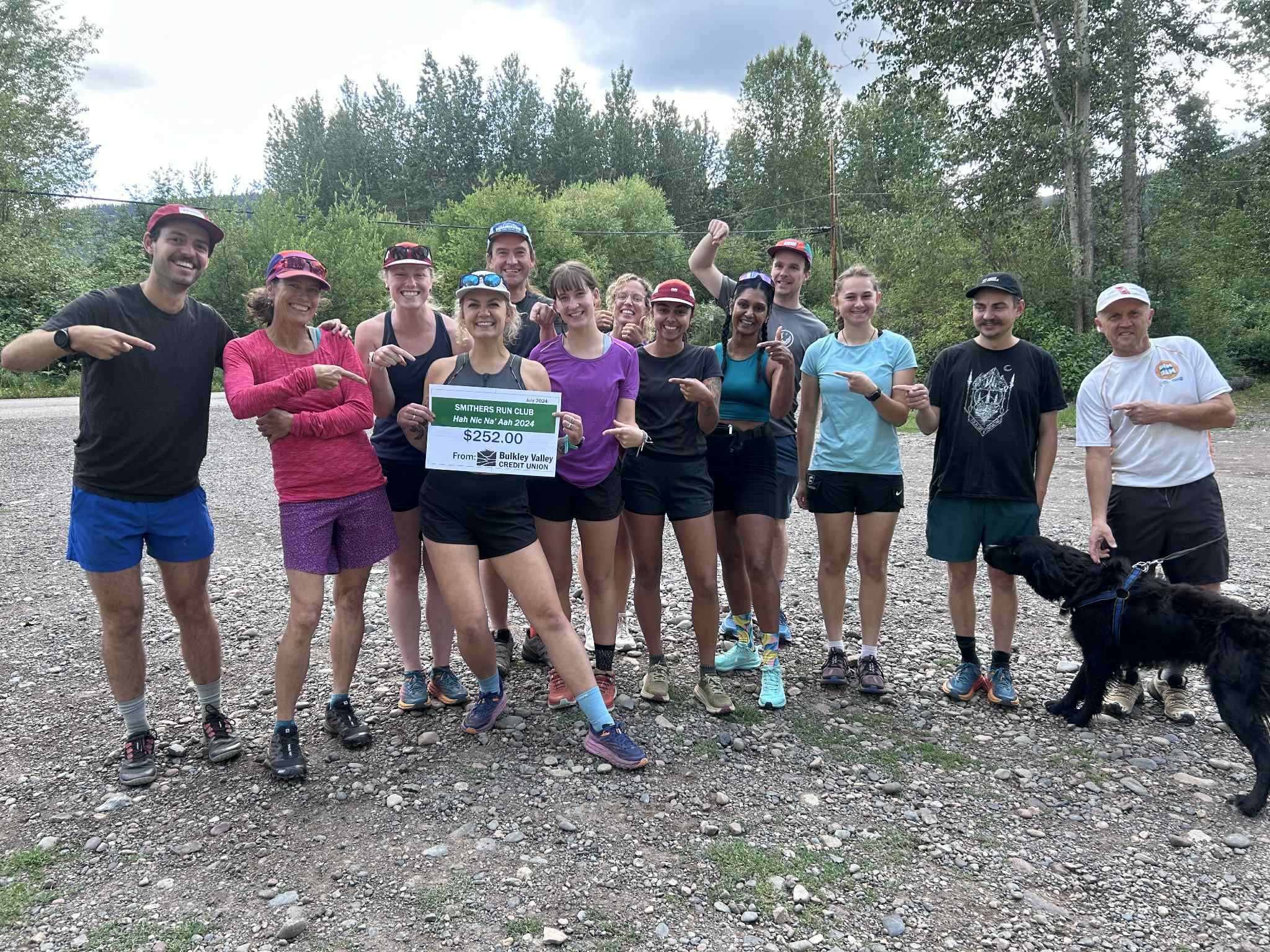 Group of people, mostly women in athletic gear, standing outdoors on a gravel surface with trees in the background, holding a large check for $252 from Smithers Run Club, celebrating a running event.
