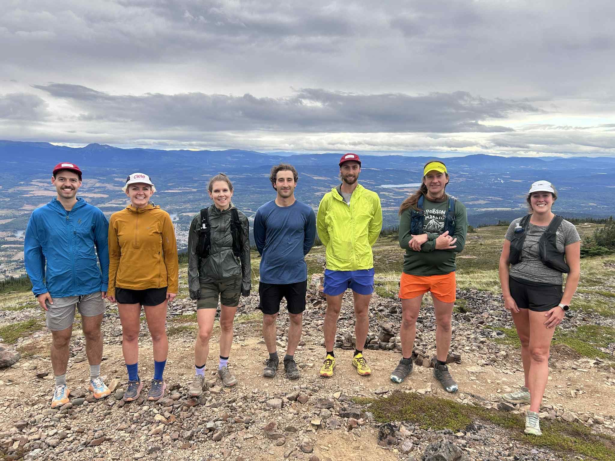 Group of seven trail runners standing on a rocky mountain summit with a view of mountains and valley in the background