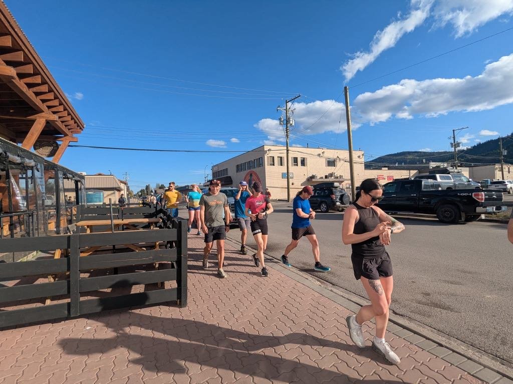 A group of people jogging on a city sidewalk on a sunny day with a blue sky and some clouds.