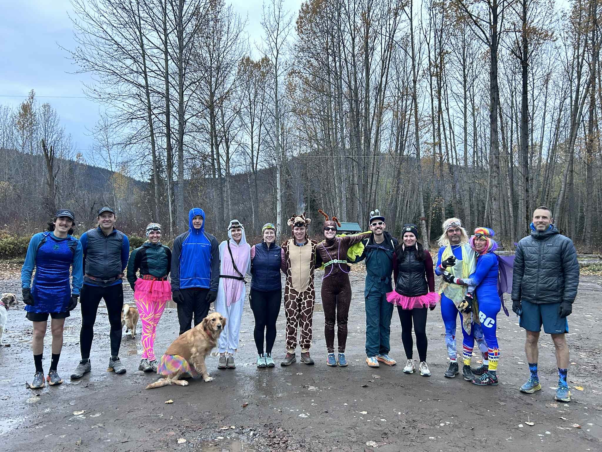 Group of people dressed in colorful athletic and costume outfits standing outdoors in a wooded area, some with dogs.