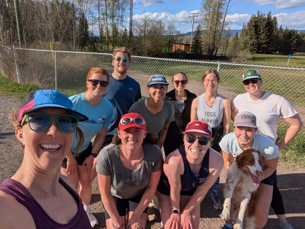 Group of ten smiling women and one dog outdoors on a sunny day, standing behind a chain-link fence with trees and a horse in the background.