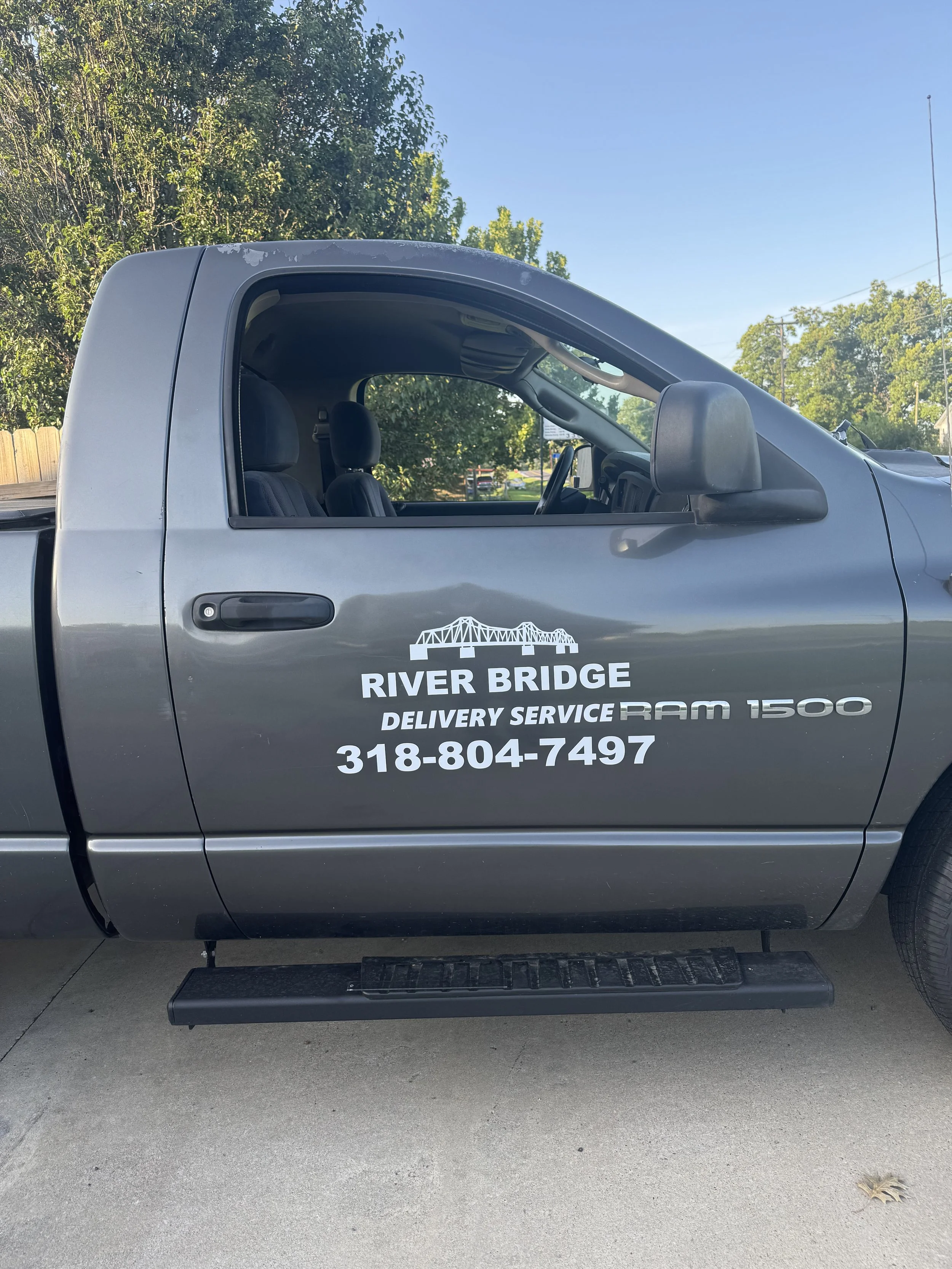 Side view of a gray pickup truck with business advertising on the door for River Bridge Delivery Service, including a bridge graphic, phone number, and vehicle model Ram 1500.