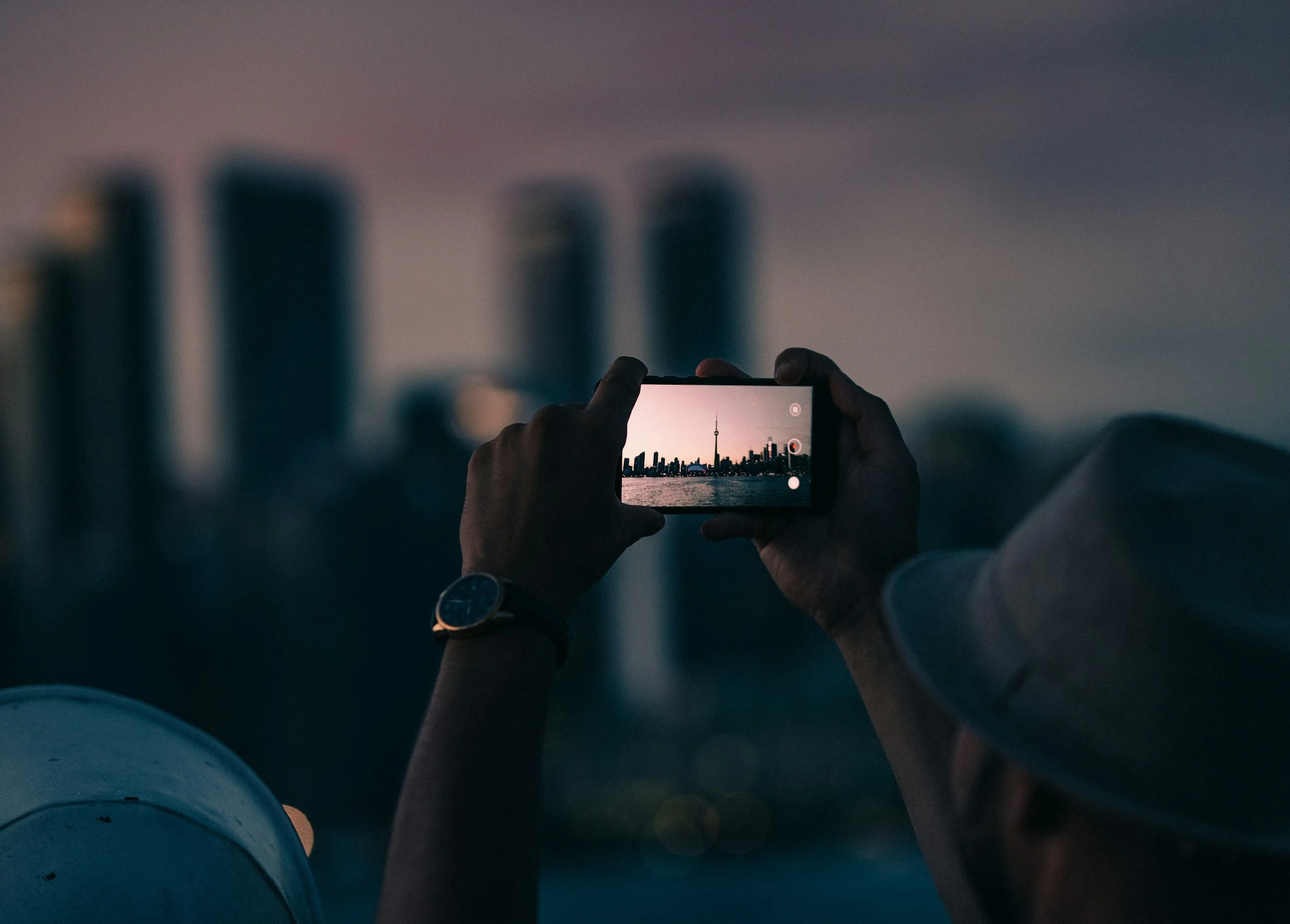 Person taking a photo of a city skyline with a smartphone at dusk, with some buildings blurred in the background.