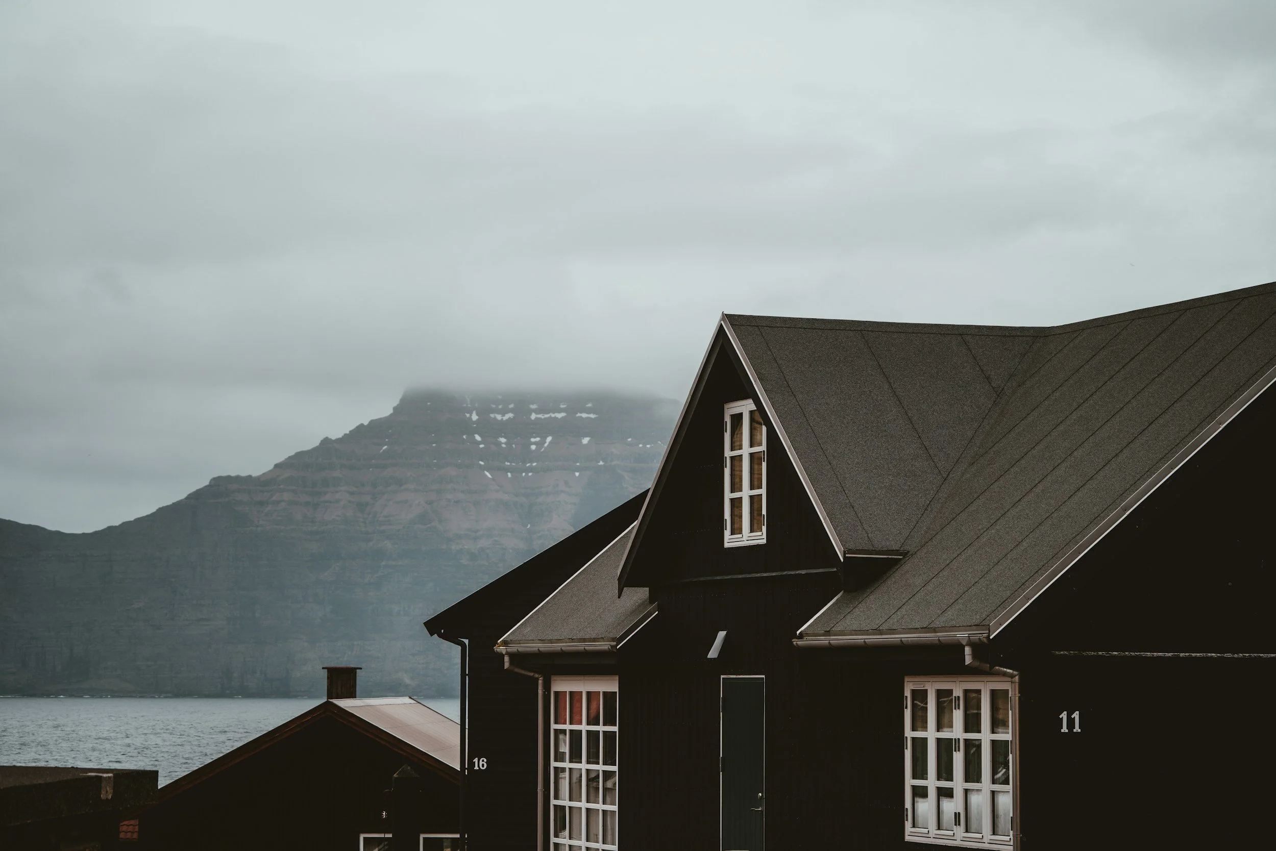 A black house with a steep roof and large windows, near water with mountains in the background under a cloudy sky.