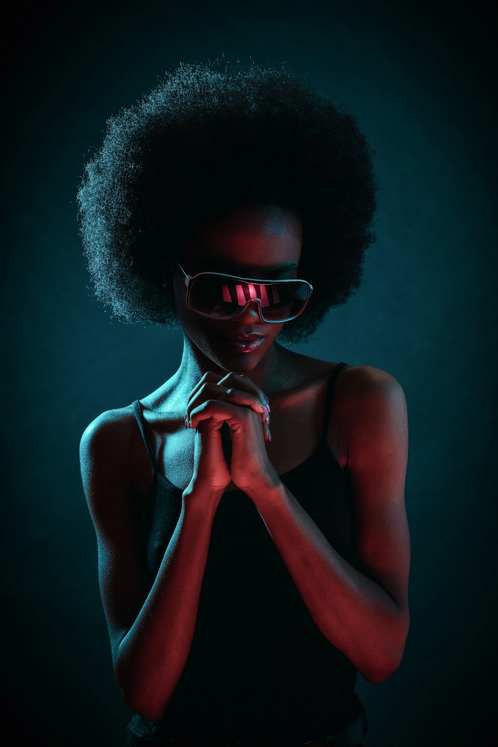 A woman with dark skin and an afro hairstyle wearing reflective sunglasses and a black tank top, illuminated by dim blue and red lighting.