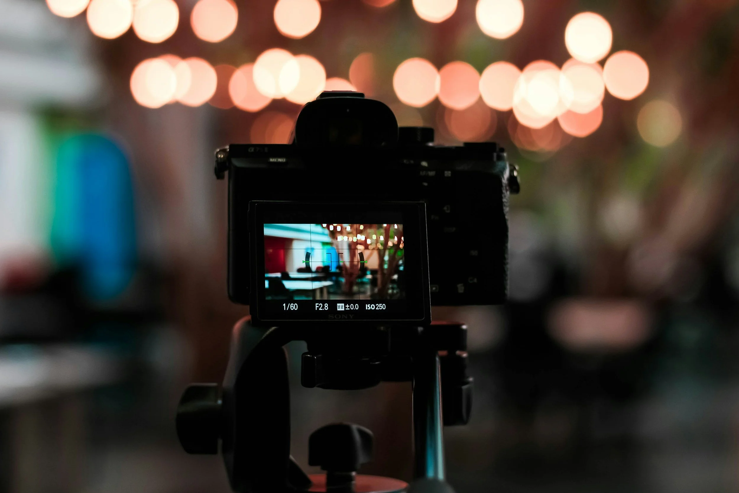 A camera on a tripod capturing an indoor scene with blurred string lights, a tree, and colorful decor in the background.