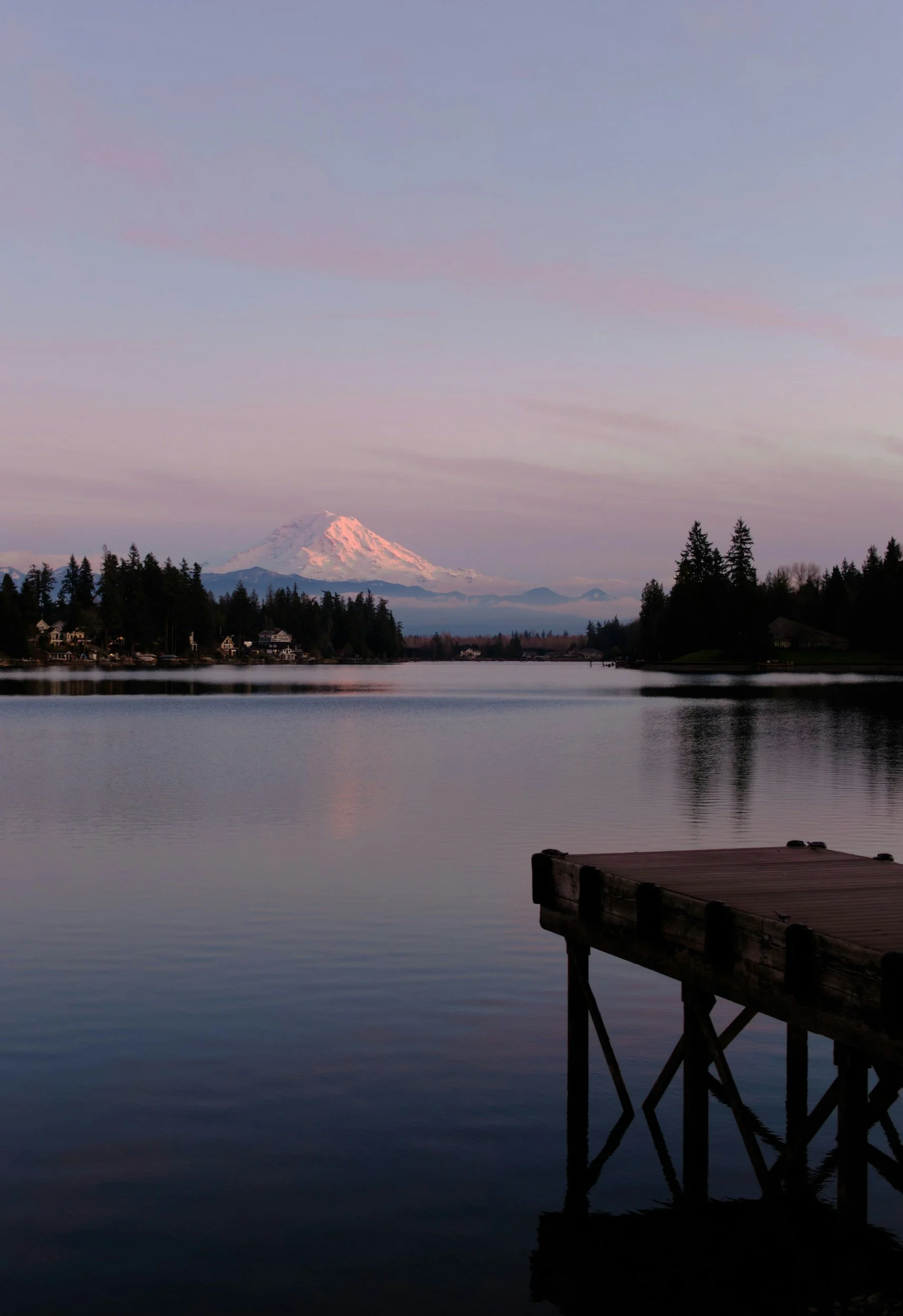 Snow-capped mountain in the distance across a calm body of water with a wooden dock in the foreground and a few houses and trees along the shore.
