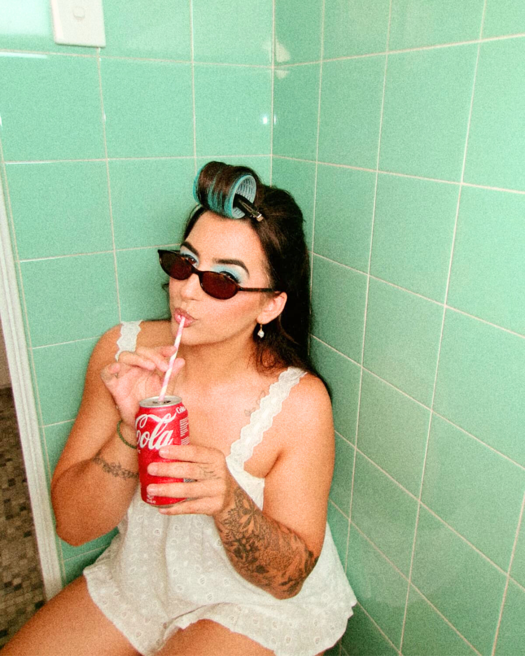 A woman with blue eyelashes and eye makeup wearing sunglasses, sitting in a bathroom corner with green tiled wall, drinking from a Coca-Cola can with a straw, wearing white nightwear with lace straps, and hair rollers on her head.