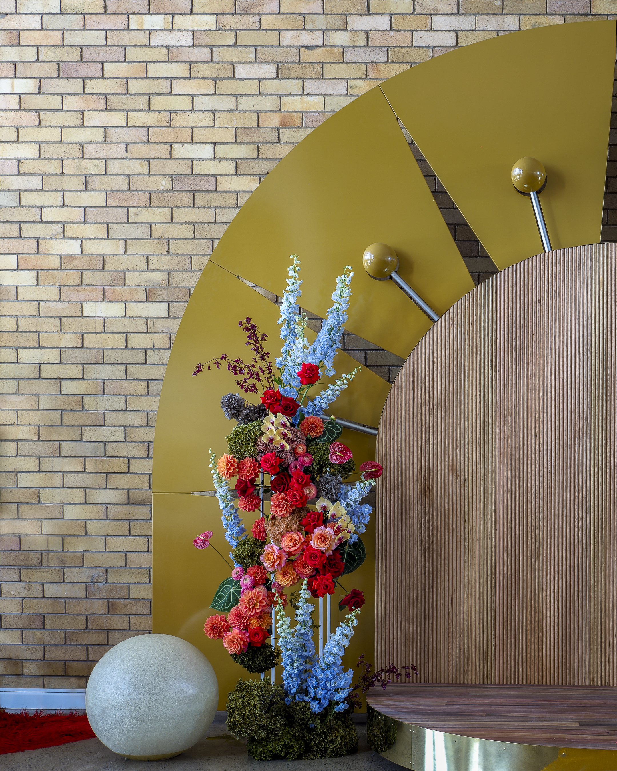 An abstract art installation with a large yellow circular structure, vertical ribbed wooden panel, and colorful flower arrangement in front of a brick wall.