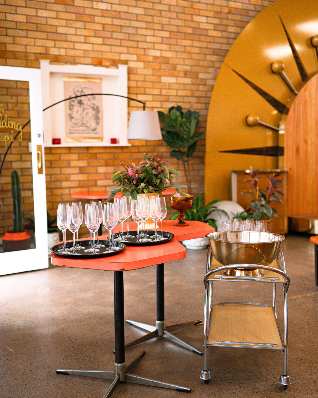 A decorated indoor space with a coral-colored table holding multiple empty champagne glasses on trays, a centerpiece with pink flowers, and a metal bowl on a trolly. In the background are plants, a brick wall, framed artwork, a white shelf, and a lar