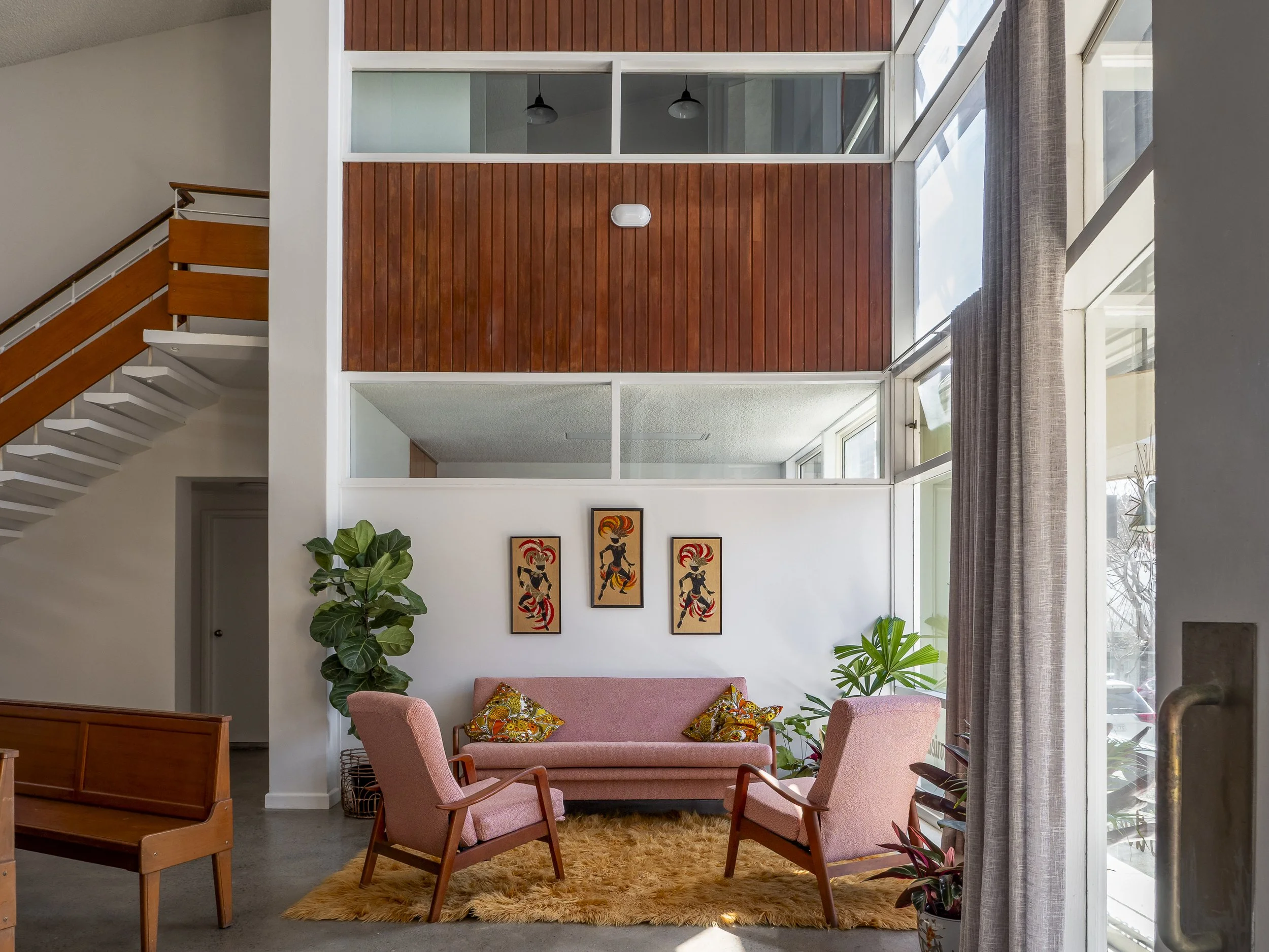 A cozy living room with a pink sofa and two matching armchairs arranged around a plush orange rug, decorated with colorful throw pillows. The room features lush green potted plants and is illuminated by large windows with beige curtains. Three framed