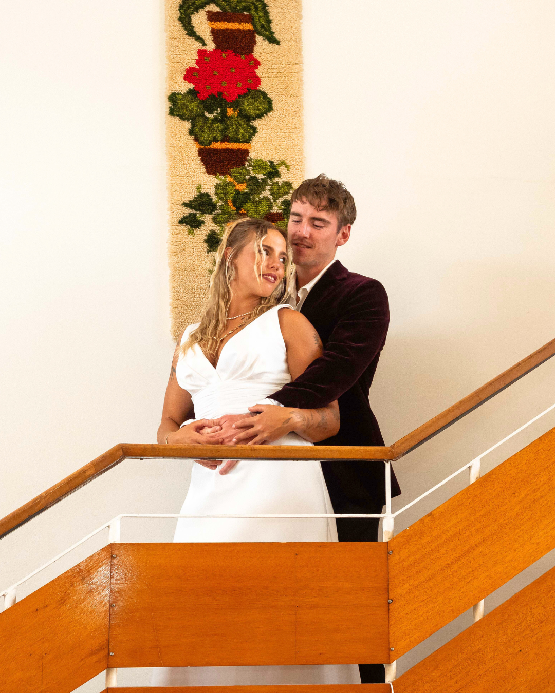 A couple, a man and a woman, standing on a staircase, embracing and smiling. The woman is wearing a white dress, and the man is dressed in a dark suit jacket. Behind them is a decorative wall hanging featuring a potted plant.