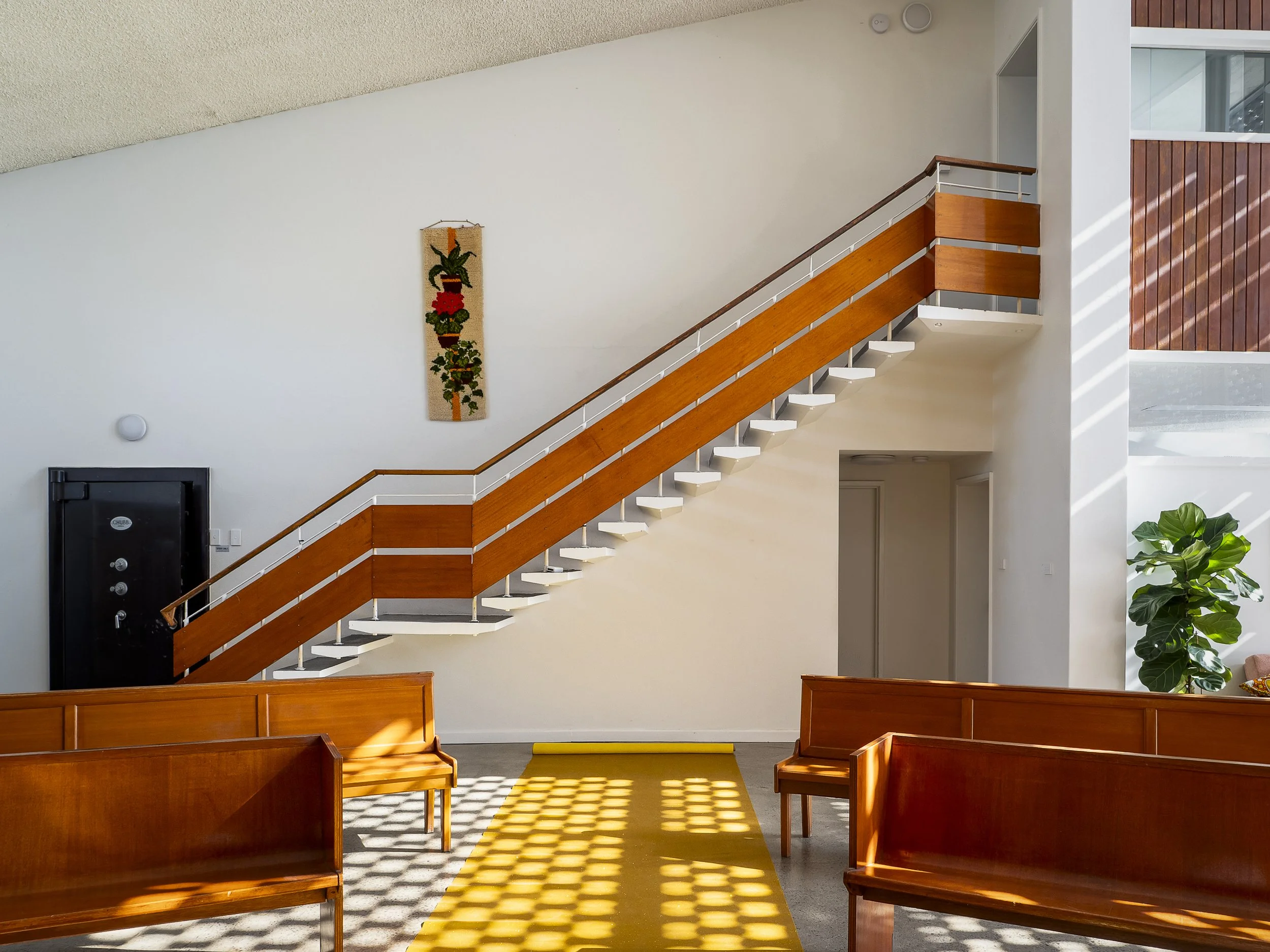 Interior view of a building lobby with wooden benches, a yellow carpet, stairs with wooden railings leading up, white walls, a wall hanging plant tapestry, a large leafy plant, and natural light streaming in.