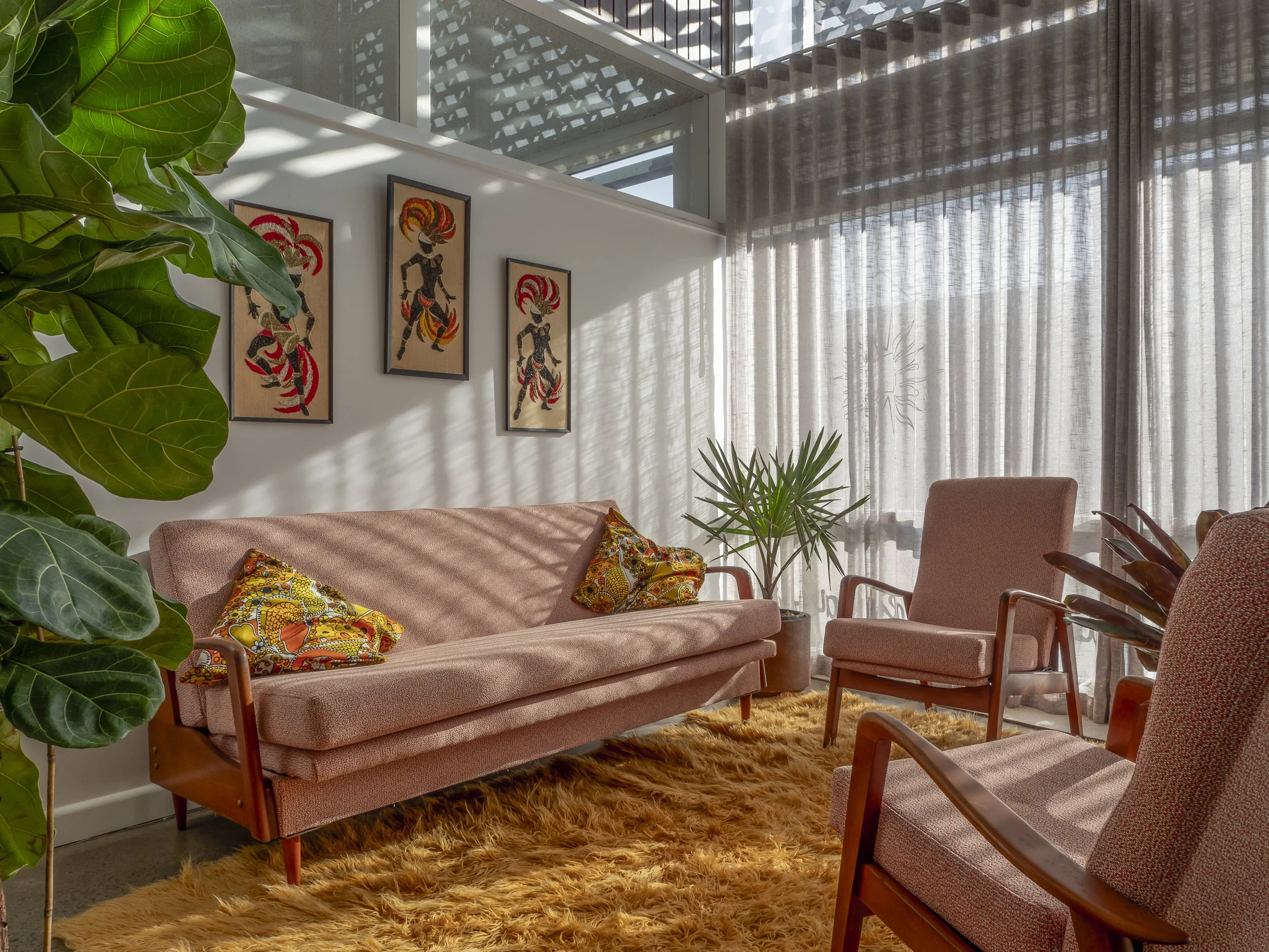 A cozy living room with pink upholstered furniture, including a sofa and armchairs, decorated with colorful pillows, and a fluffy yellow rug. There are several green plants and framed artwork on the wall, with a large window covered by beige curtains