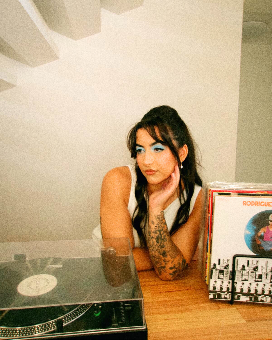 A woman with dark hair and tattoos resting her chin on her hand at a table, with a vinyl record player and a collection of records nearby.
