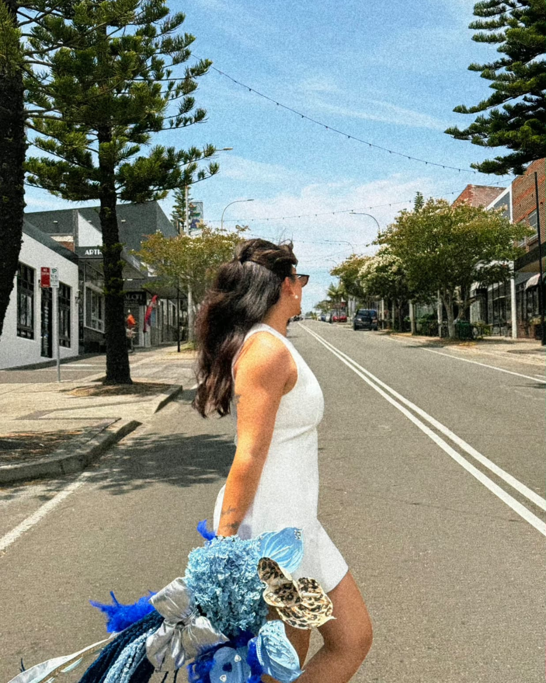 A woman with long dark hair, wearing sunglasses and a white dress, crossing an empty street on a sunny day, holding a large bouquet of blue, white, and animal print fabric flowers.