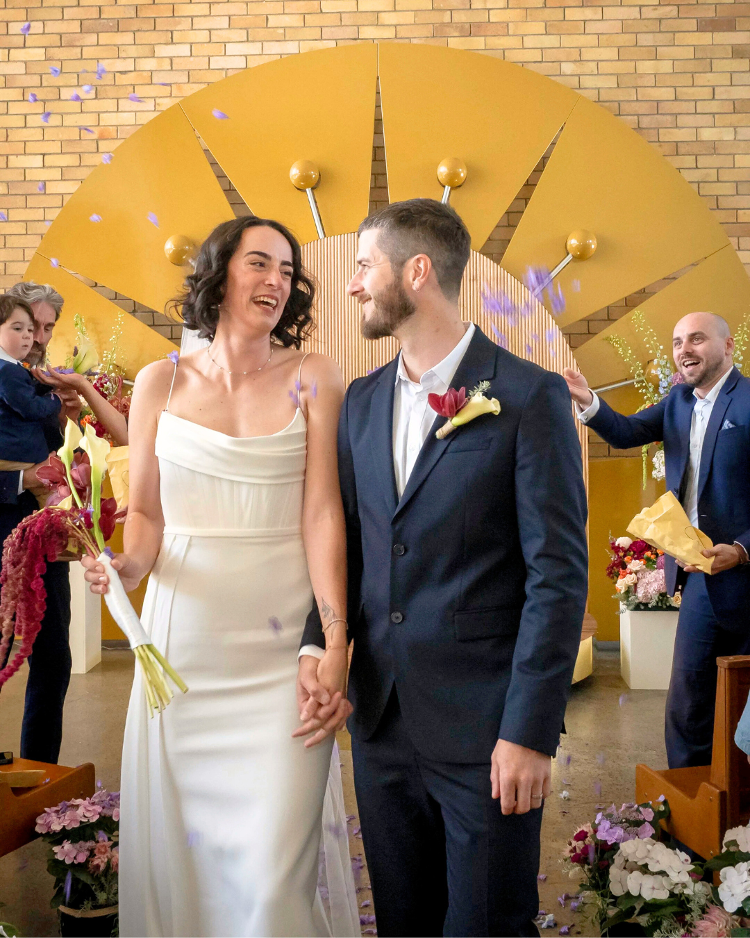 A bride and groom holding hands and smiling at each other during their wedding ceremony, standing in front of a large, gold, clock-like decorative backdrop, with friends and family celebrating around them, releasing purple flower petals.