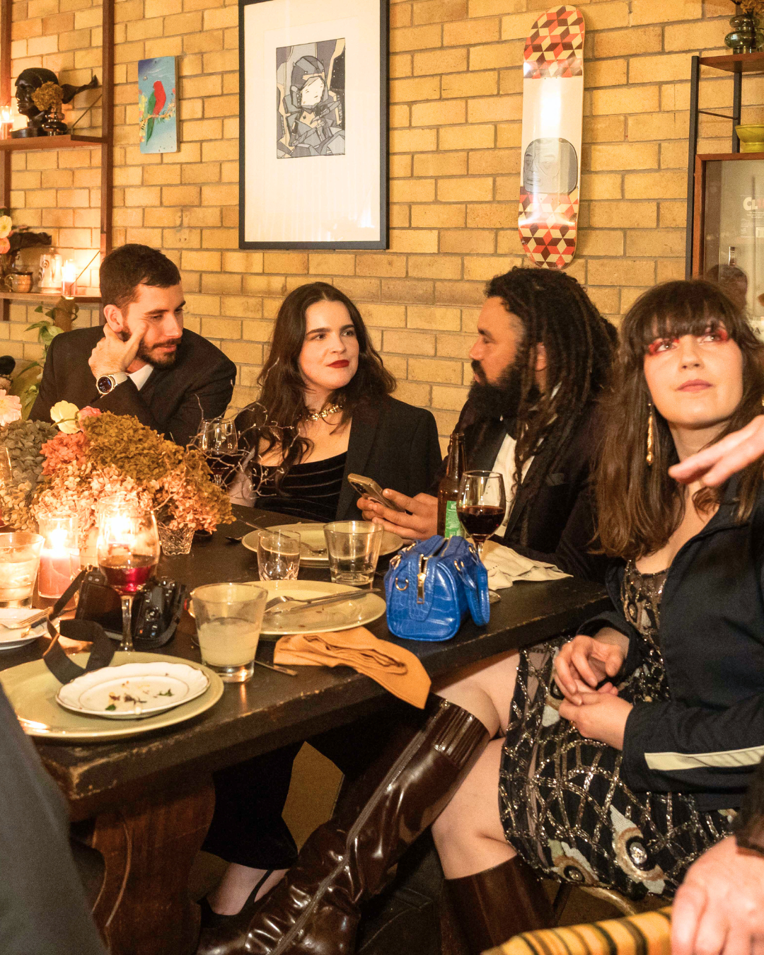 People gathered around a dinner table in a cozy, decorated restaurant with brick walls, engaged in conversation, with food, drinks, and flowers on the table.
