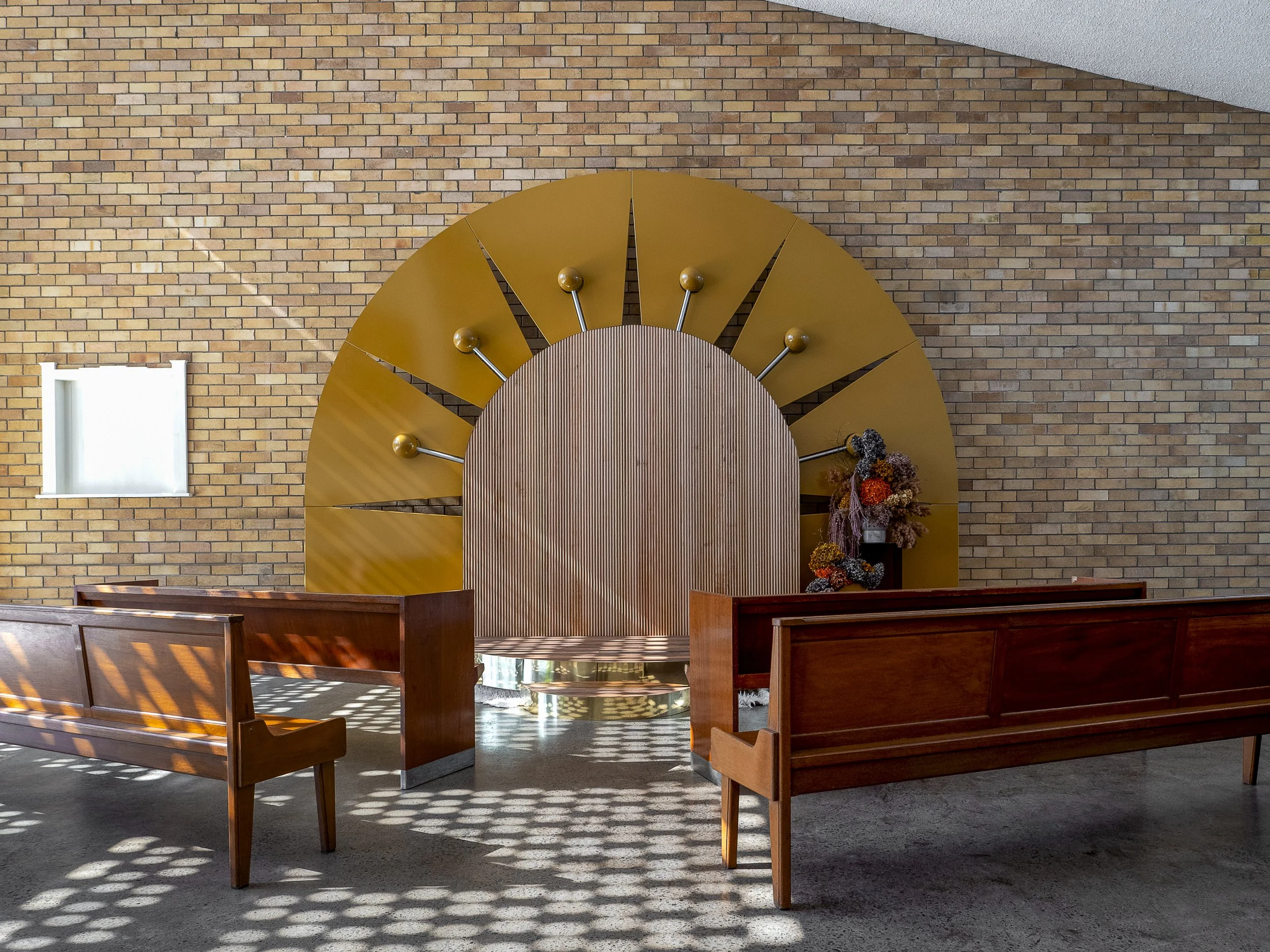 Interior of a church or chapel with a wooden altar against a brick wall, decorated with flowers and sunlit wooden benches.