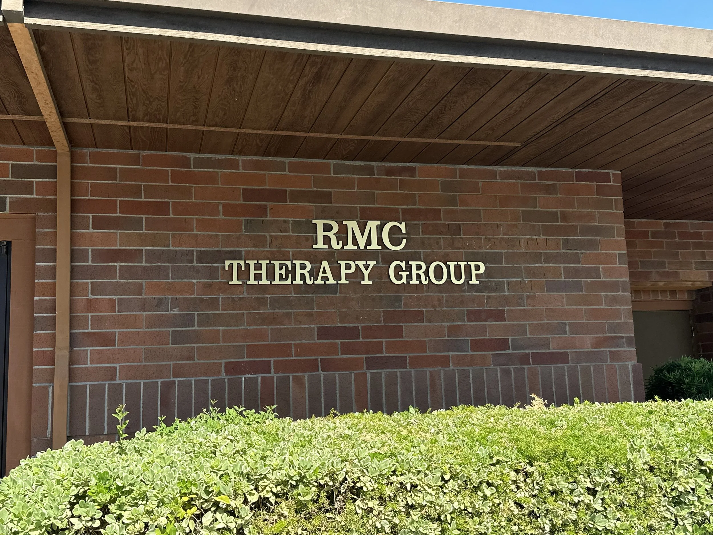 Sign on brick wall reading 'RMC Therapy Group' outside a building with green bushes in front.