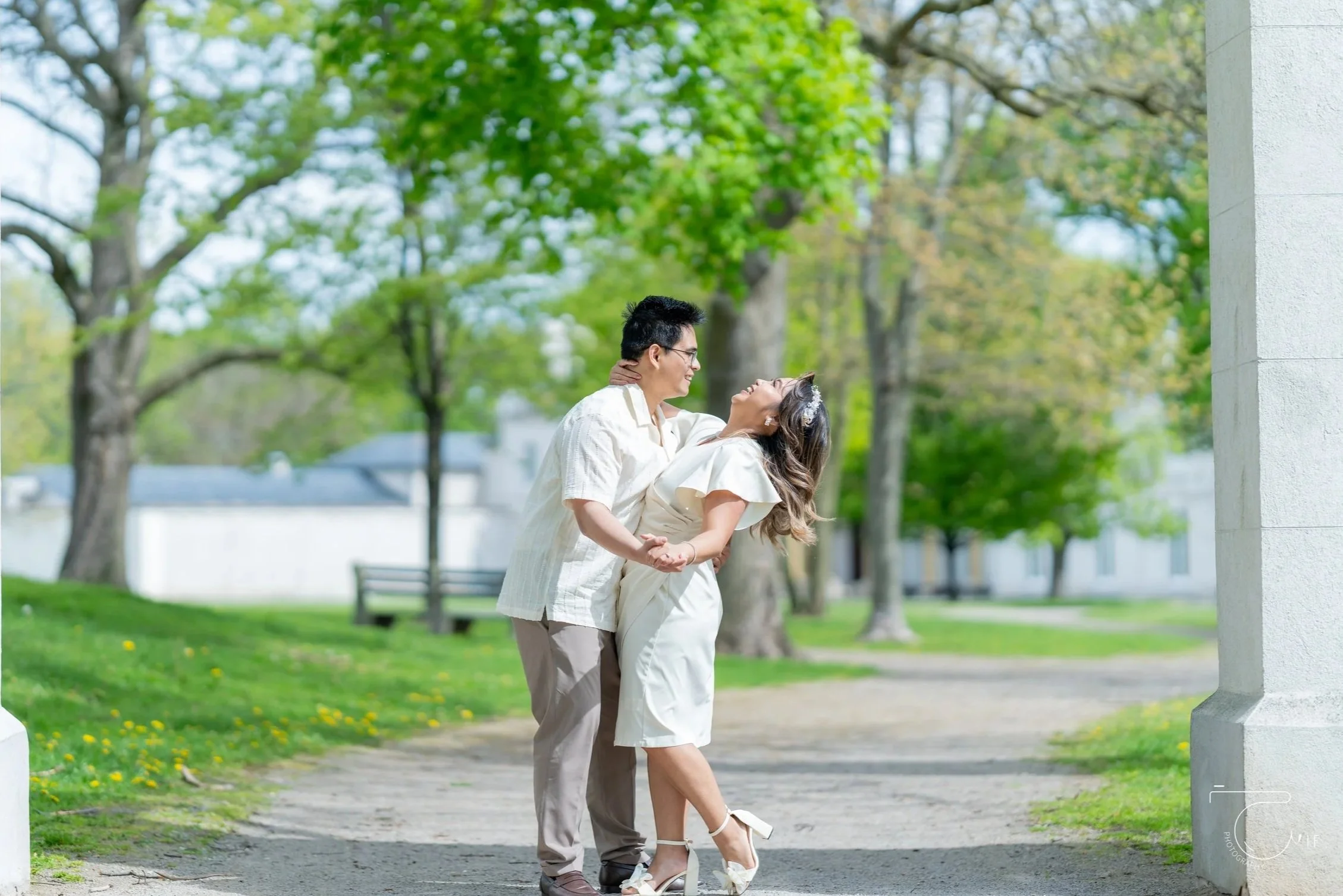 A couple dancing and laughing outdoors in a park with green trees and sunlight.