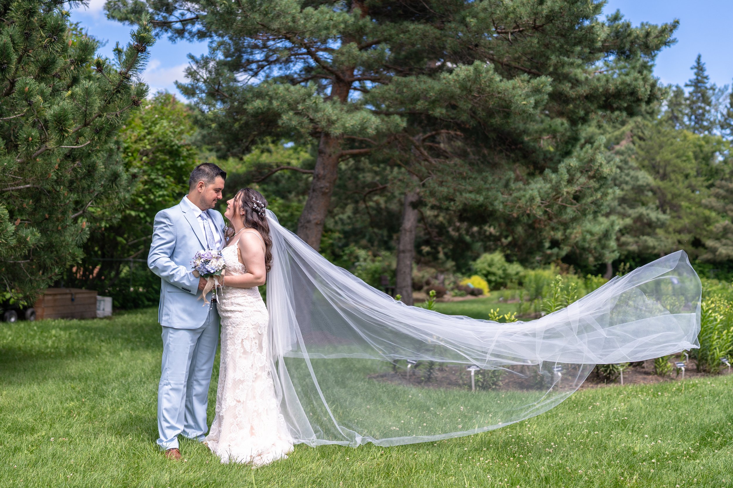 A bride and groom are standing close together on a lush green lawn, holding hands, with the bride wearing a long, flowing veil and a white wedding dress, and the groom dressed in a light blue suit. They are gazing into each other's eyes, surrounded by tall trees and a garden.