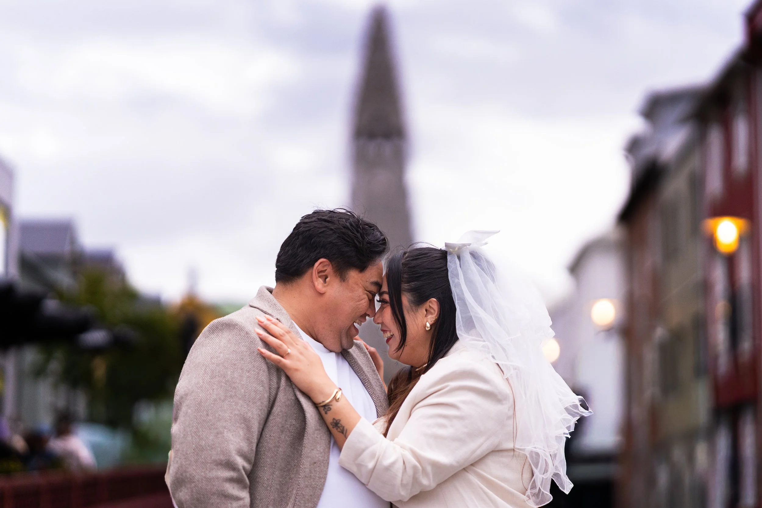 A couple smiling and touching foreheads on a city street with a clock tower in the background.