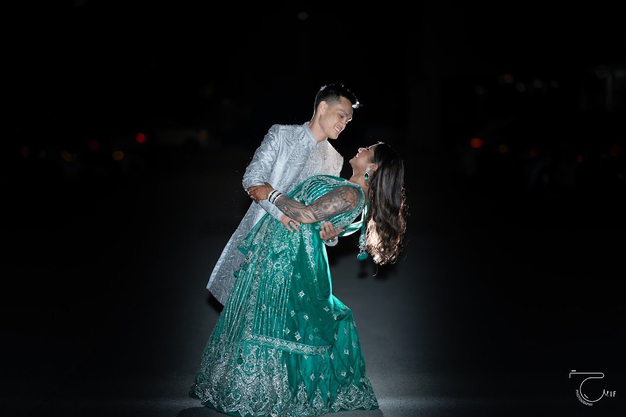 A couple dancing together at night, with the man wearing a traditional silver outfit and the woman in a green saree with intricate silver embellishments.