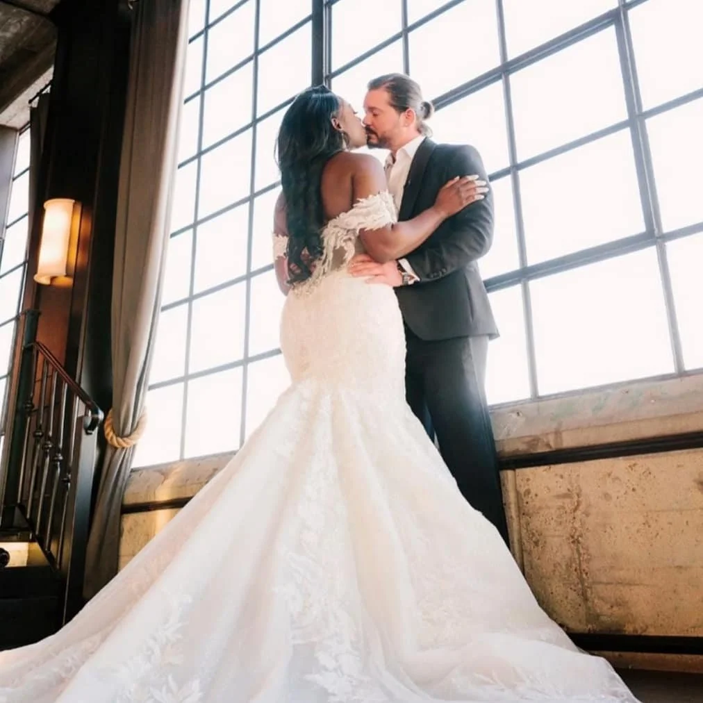 A bride and groom sharing a kiss in a wedding dress and suit inside a room with large windows and natural light.