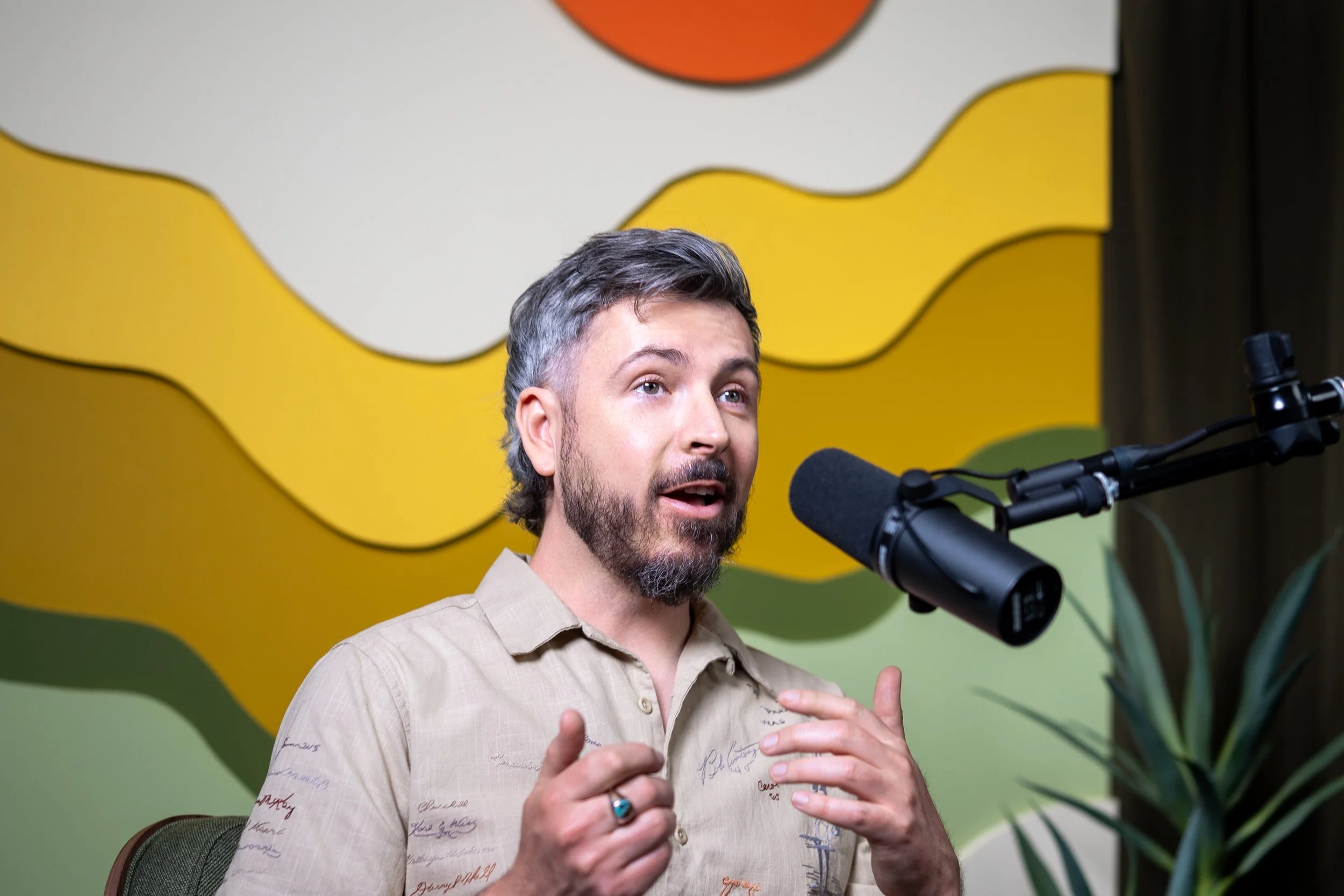A man speaking into a microphone with a colorful abstract background and a plant visible to the right.