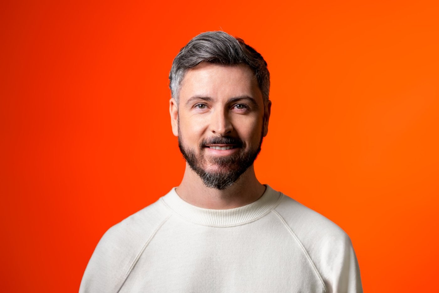 A smiling man with a beard and short hair wearing a light-colored crew neck sweater, standing against an orange gradient background.