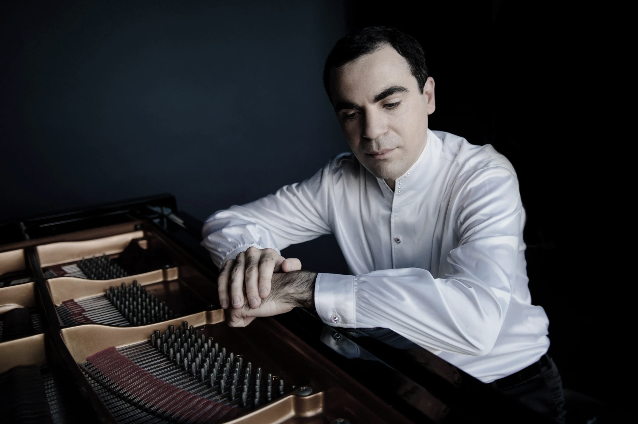 A man dressed in a white shirt with cufflinks is sitting at a grand piano, gently resting his hand on another person's head. The interior of the piano, including strings and metal components, is visible in the foreground. The background is dark.