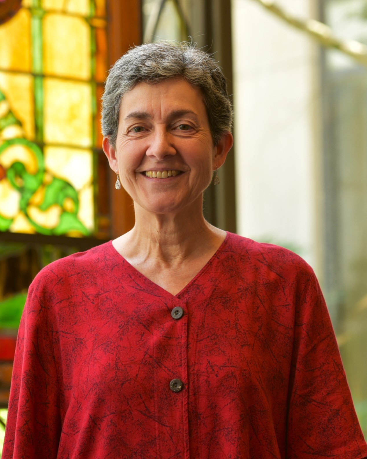 A woman with short grey hair smiling, wearing a red blouse and a naturally lit stained glass window background.