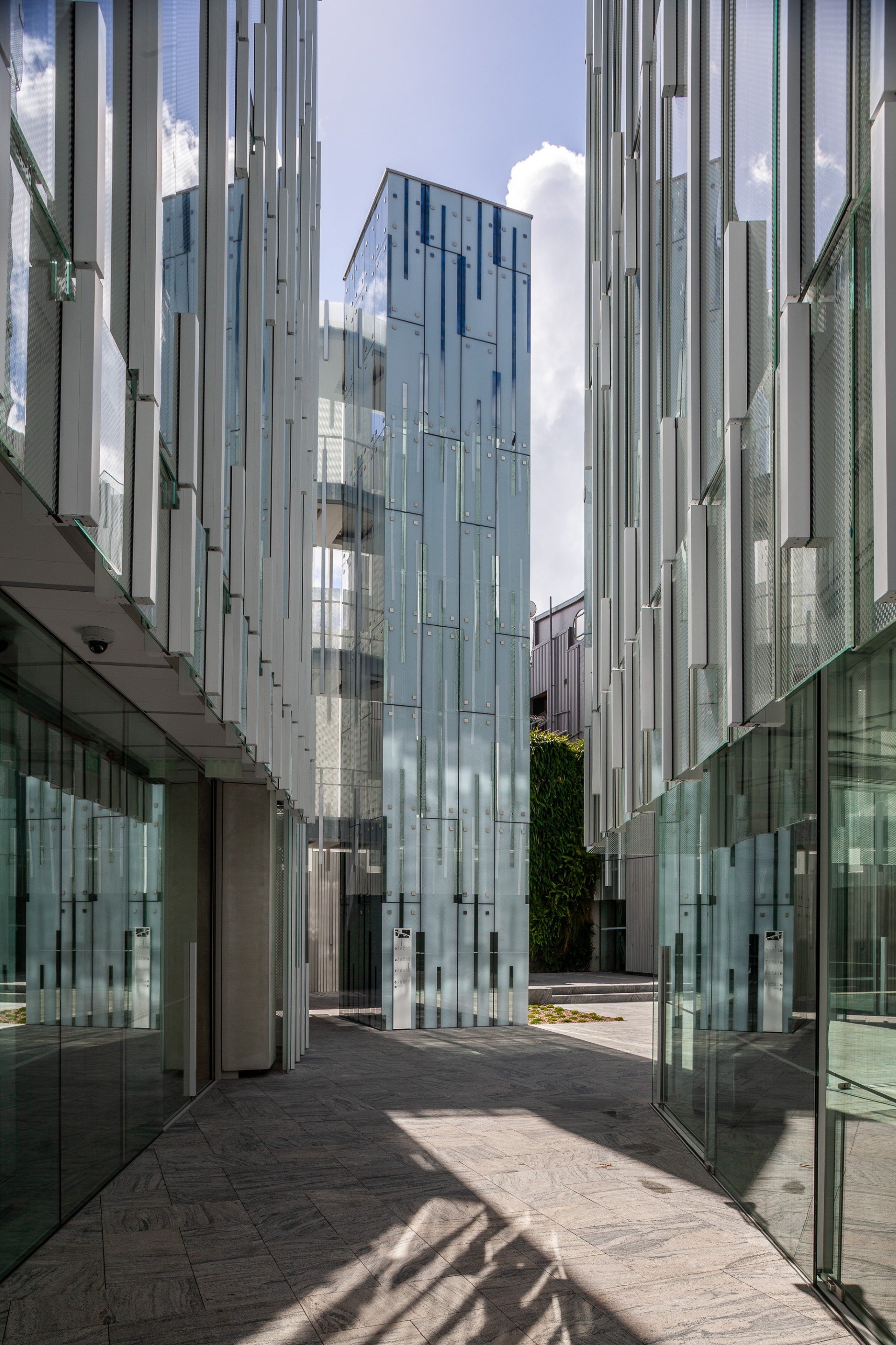 Modern office building with glass and metal facades, shadowed pathway, and blue sky with clouds.