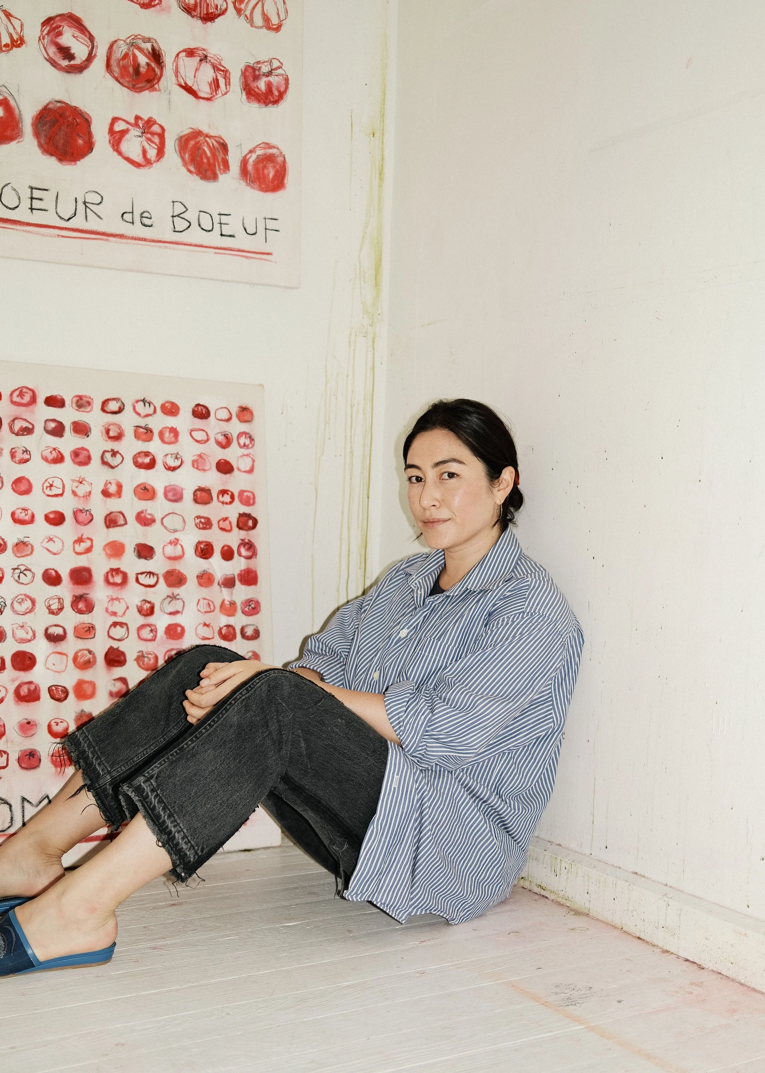A woman sitting on the floor next to a white wall, with two posters featuring red sketches of tomatoes and vegetables.