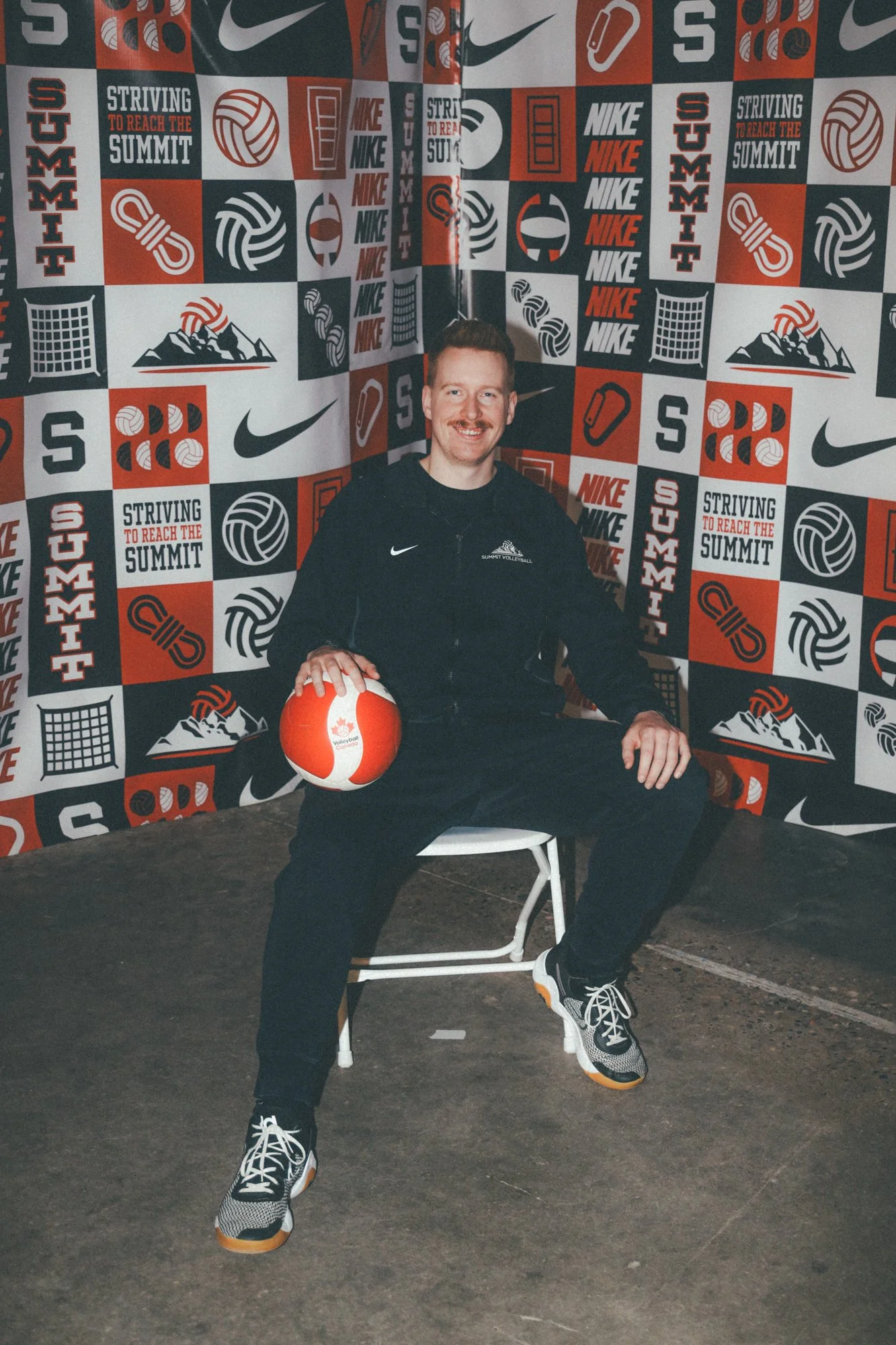 A man with a mustache and reddish hair, sitting on a white chair, holding a volleyball in his right hand. He's dressed in athletic clothing, including a black hoodie, and sits in front of a backdrop decorated with sports and motivational logos and words, such as 'Striving to Reach the Summit,' volleyball icons, mountain images, and brand logos like Nike and Nike Swoosh.