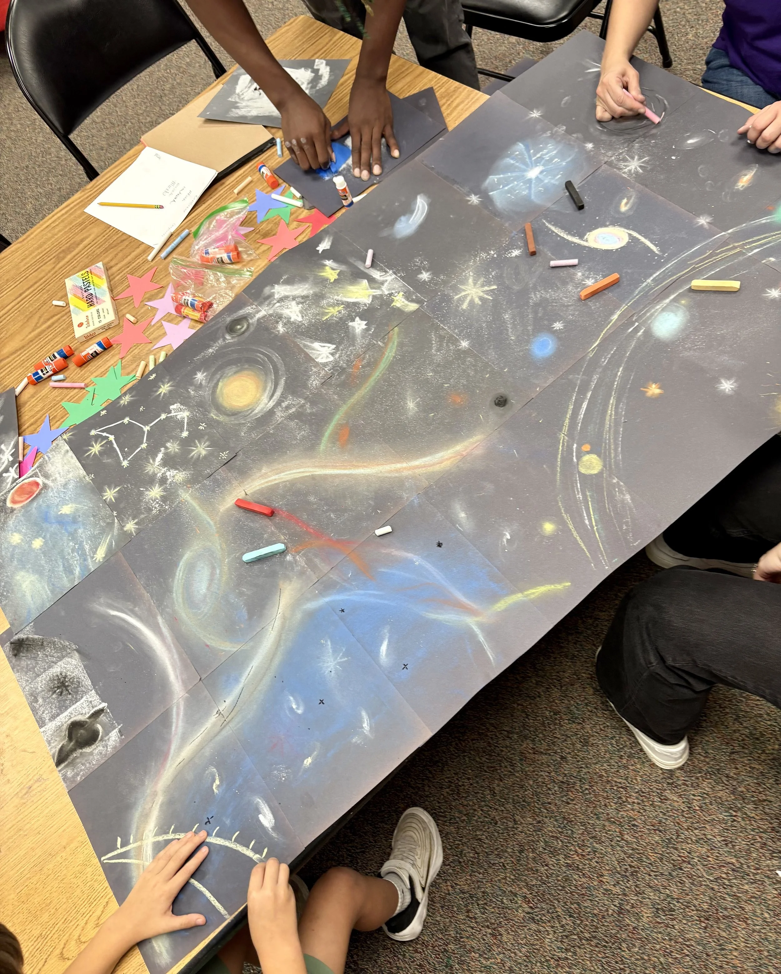 Children creating a space-themed chalk art mural on large black paper spread out on a table, with various chalks, glue sticks, and paper star cutouts nearby.