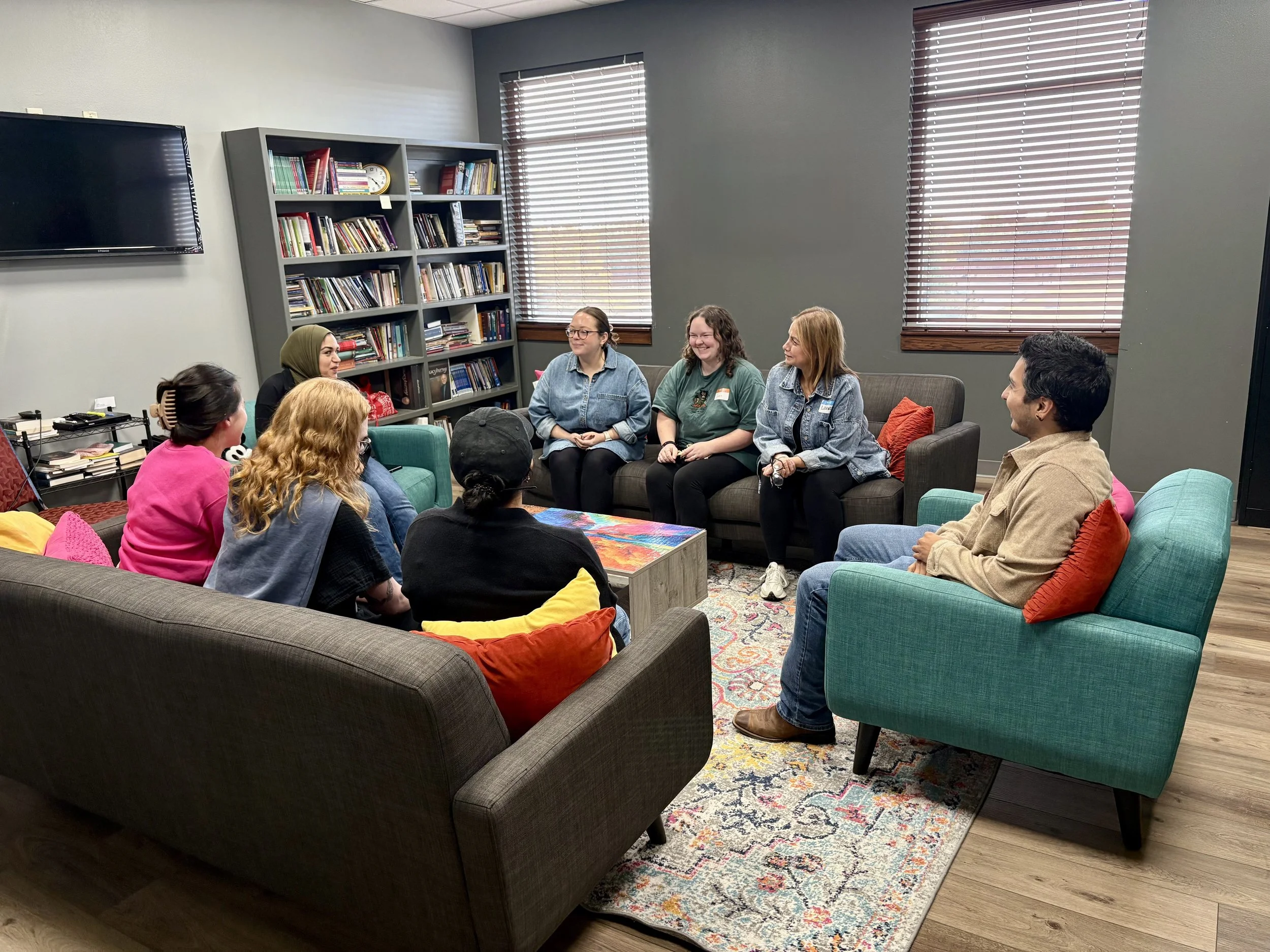 Group of eight people sitting in a semi-circle in a cozy, well-lit room with gray walls, wooden floors, and windows with blinds. There are colorful cushions on the sofas and a colorful rug on the floor, with bookshelves filled with books in the background.