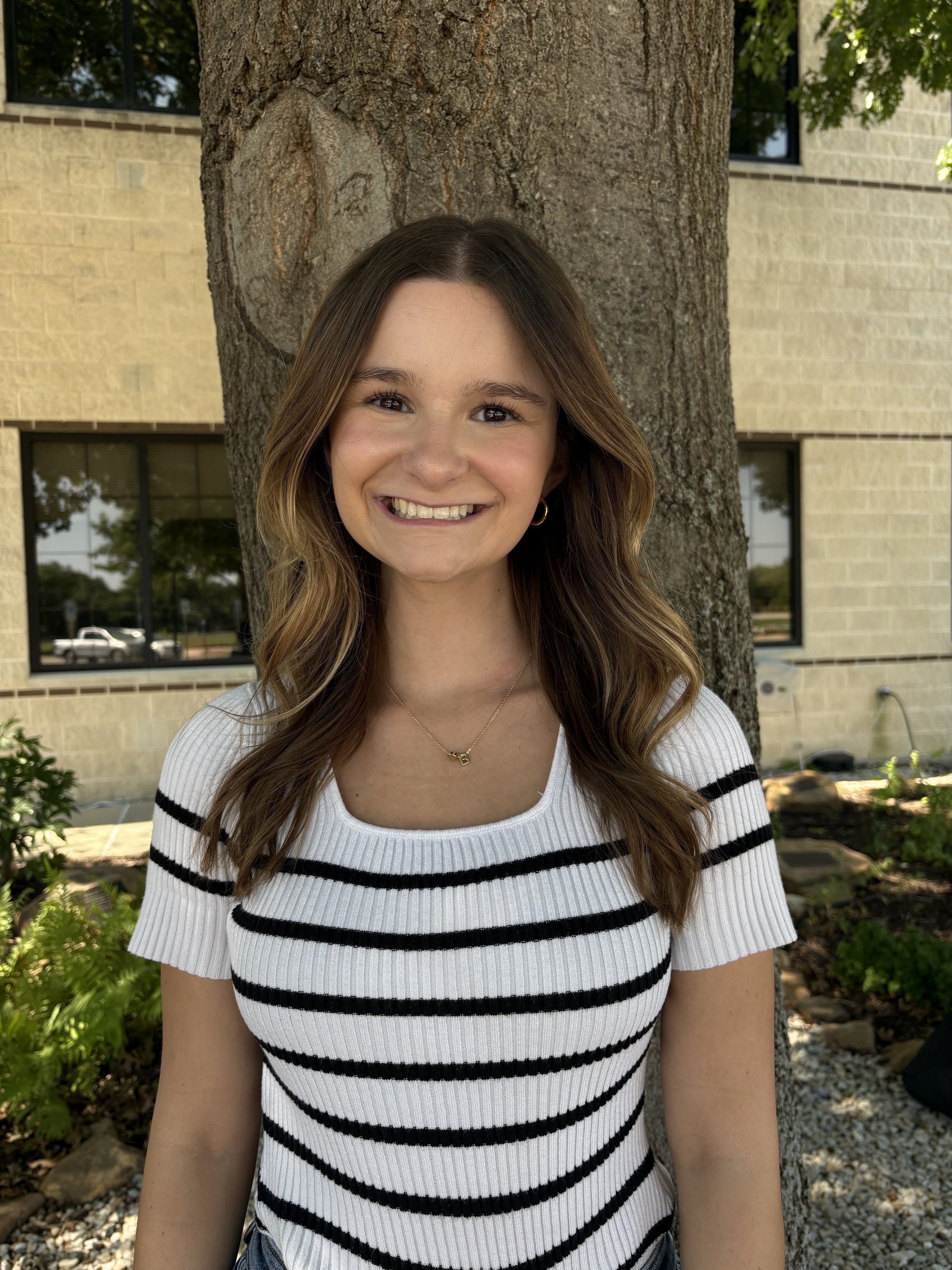 A young woman with long wavy brown hair, smiling, standing outdoors in front of a tree trunk with a brick building and windows in the background.